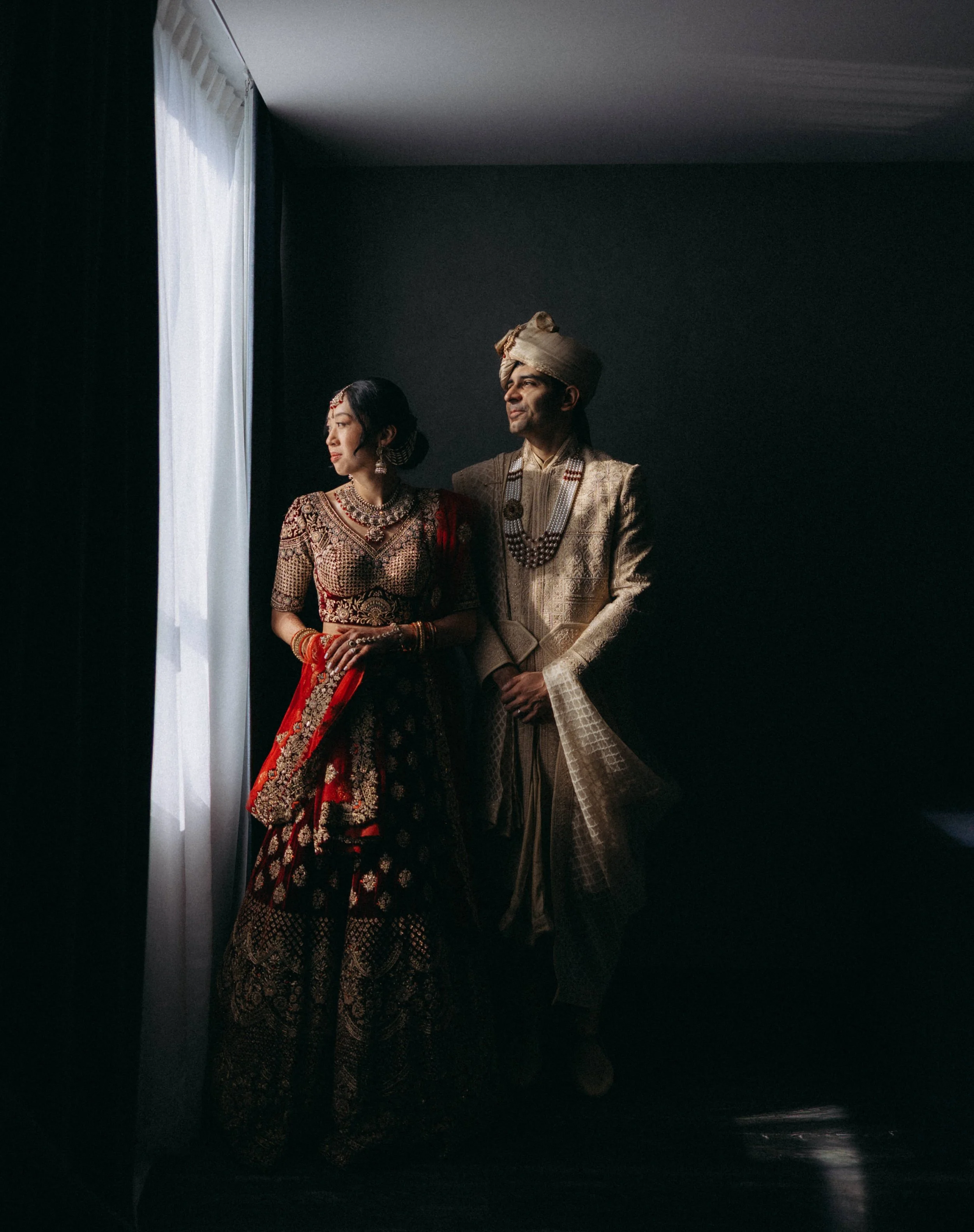 A man and a woman dressed in traditional Indian wedding attire, standing near a window with white curtains, with dark walls around them.