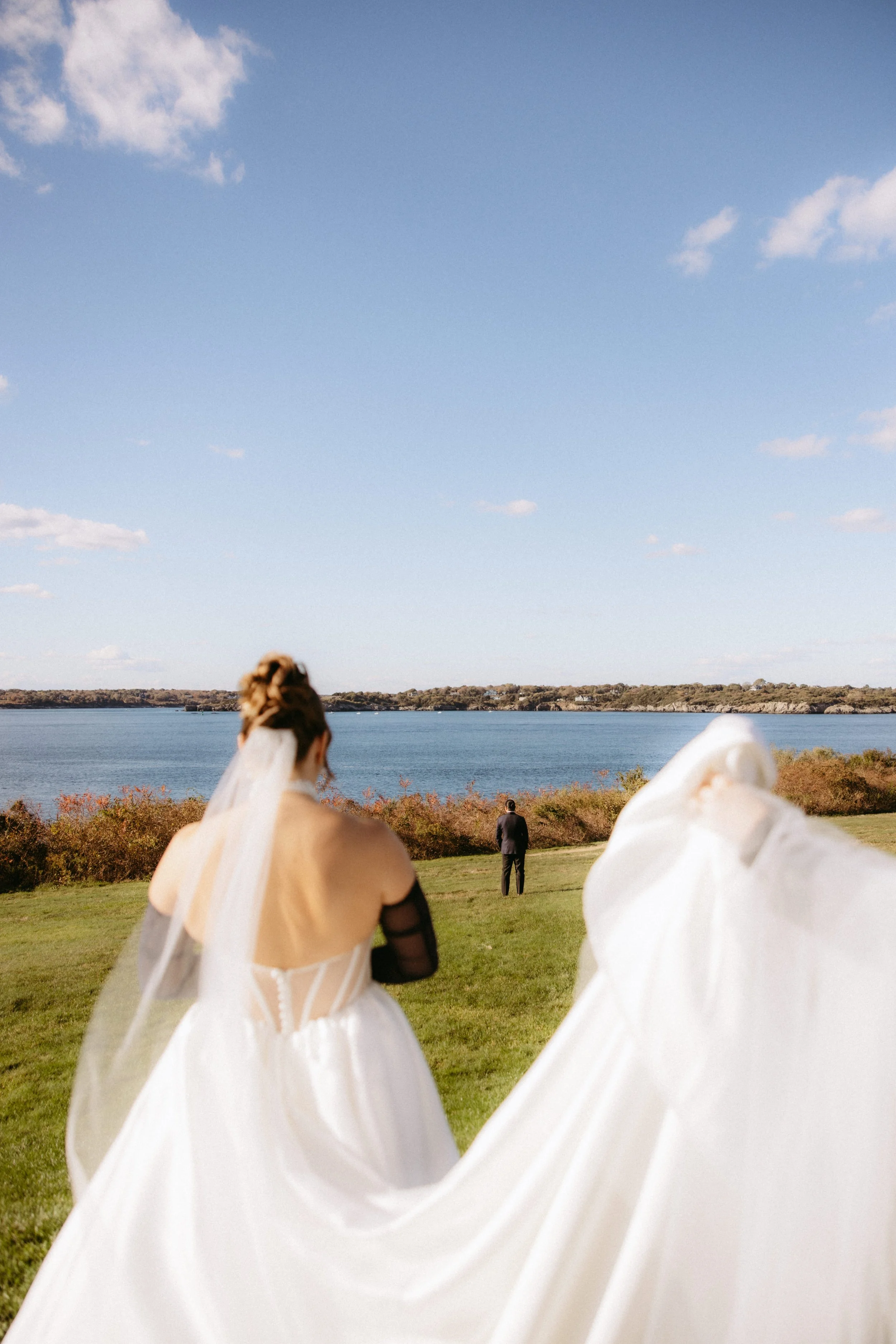 A bride and groom walking outdoors near a lake, with a woman in black and a man in a suit in the background, and a partly cloudy sky overhead.