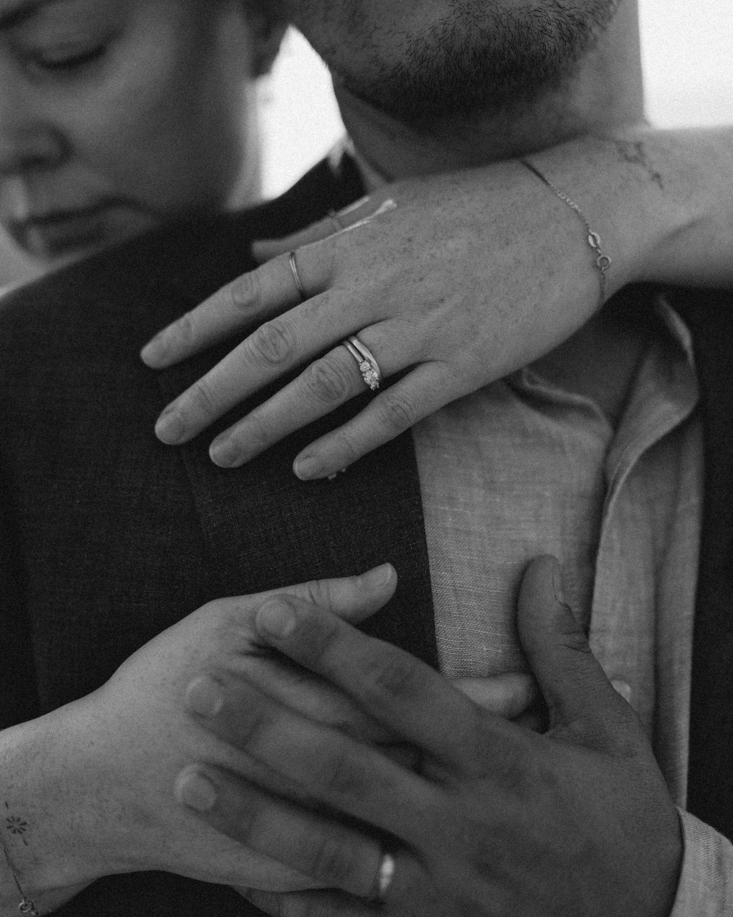 Close-up black and white photo of two people embracing. One person's hand with a ring resting on the other's shoulder. Only part of their faces and bodies are visible.