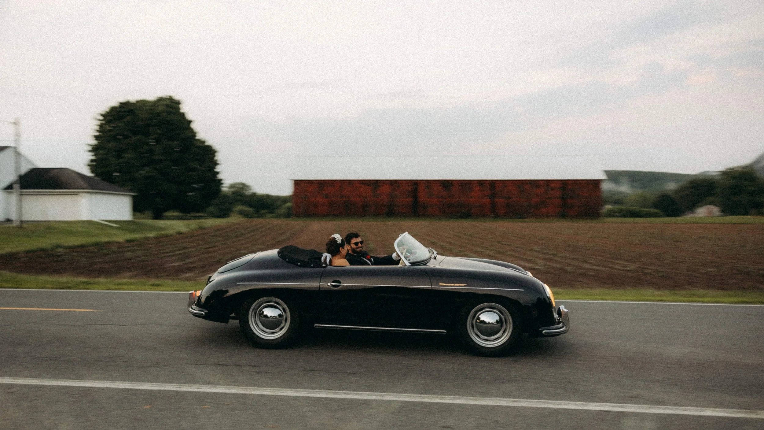 A black vintage convertible car with a man and woman inside, driving down a rural road during daytime. The woman is wearing a veil and a flower accessory, and the man is wearing sunglasses.