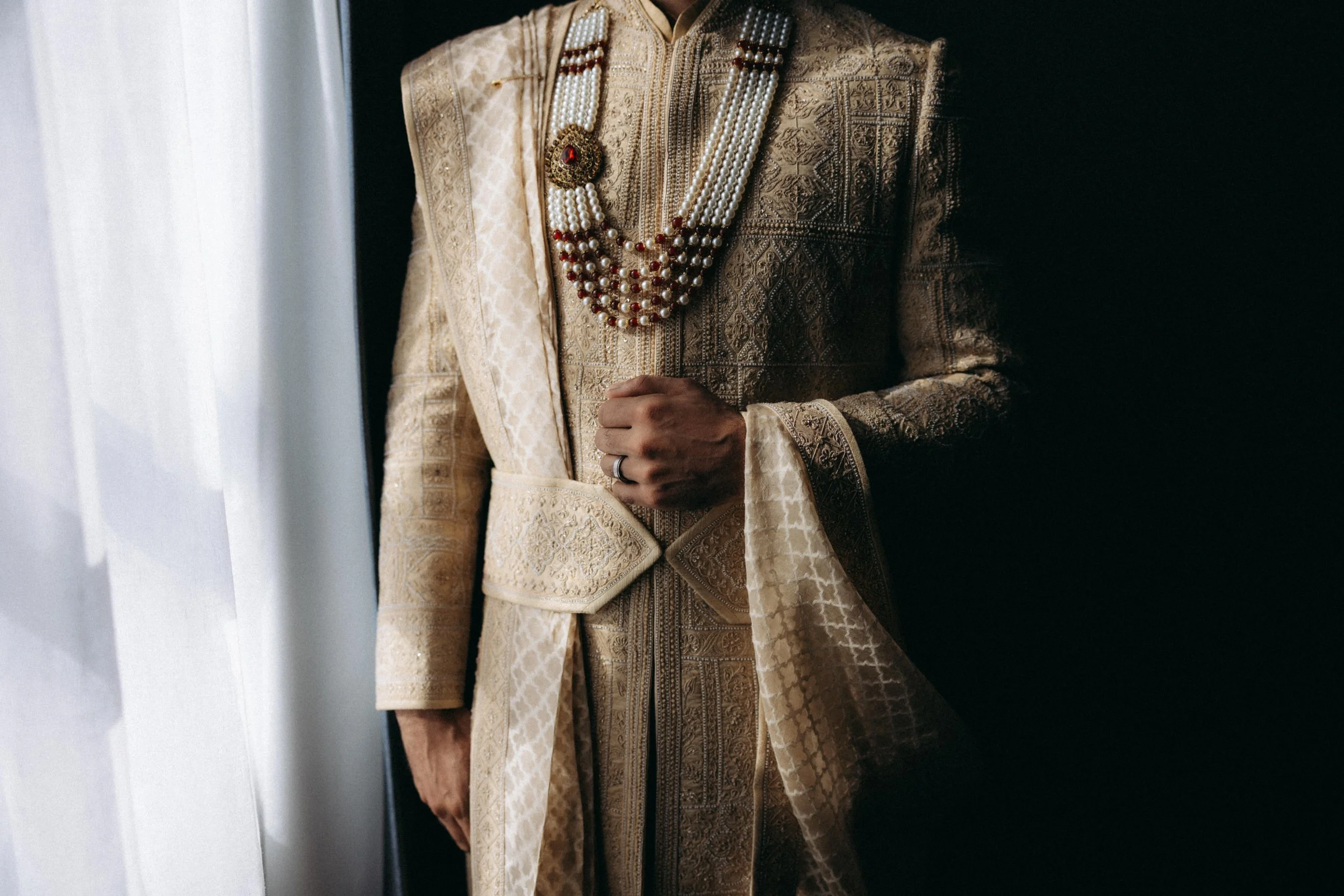 Close-up of a person wearing traditional South Asian wedding attire, including an ornate beige and gold embroidered sherwani and multiple pearl necklaces with a large red gem pendant.