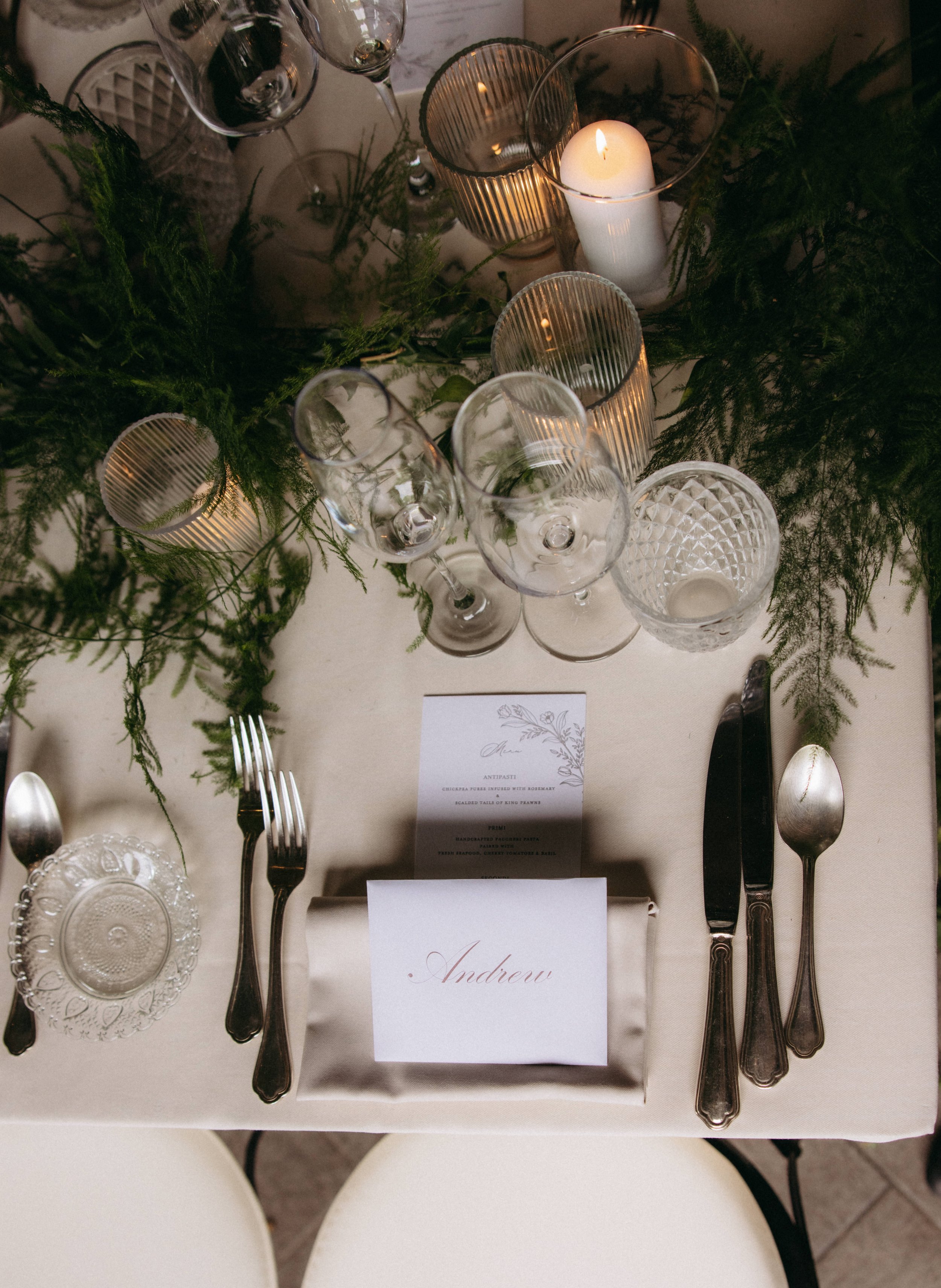 Table setting with wine glasses, candles, greenery, a menu, and a name card for Andrew.