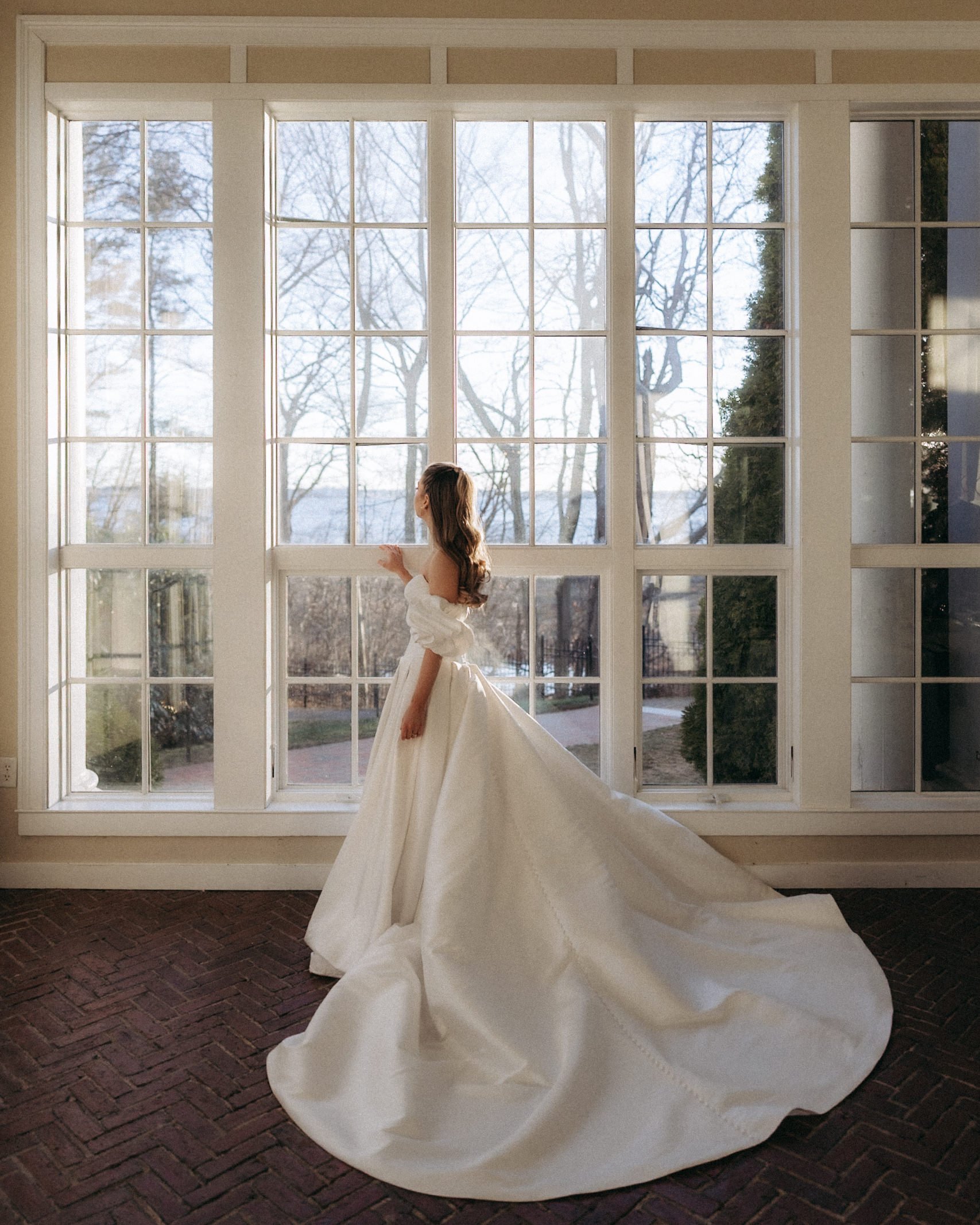 A woman in a white dress standing inside in front of a large window with multiple panes, with trees visible outside and brick flooring inside.