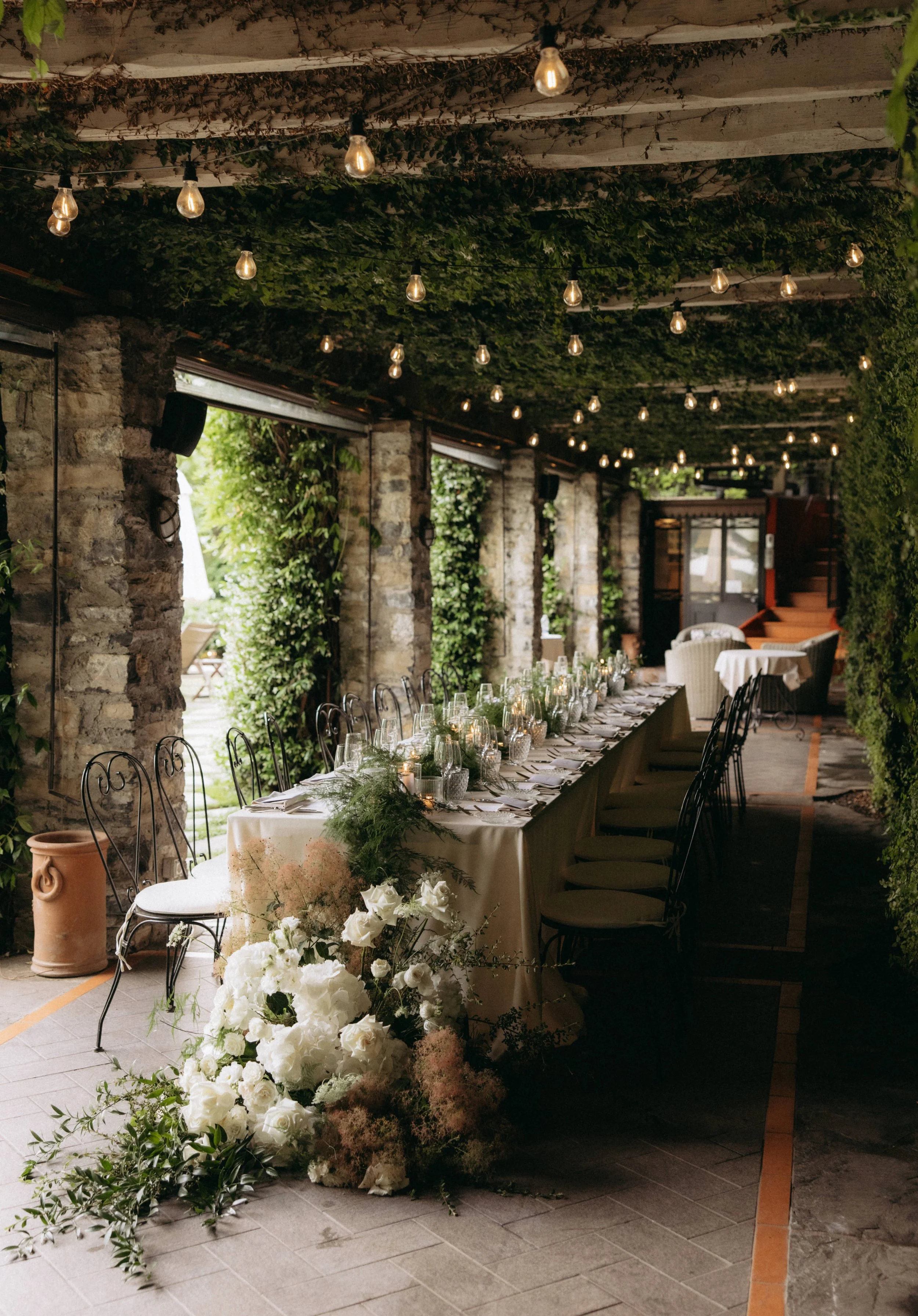 Indoor event space with a long banquet table decorated with white flowers and candles, surrounded by black chairs, under a ceiling covered with greenery and string lights.