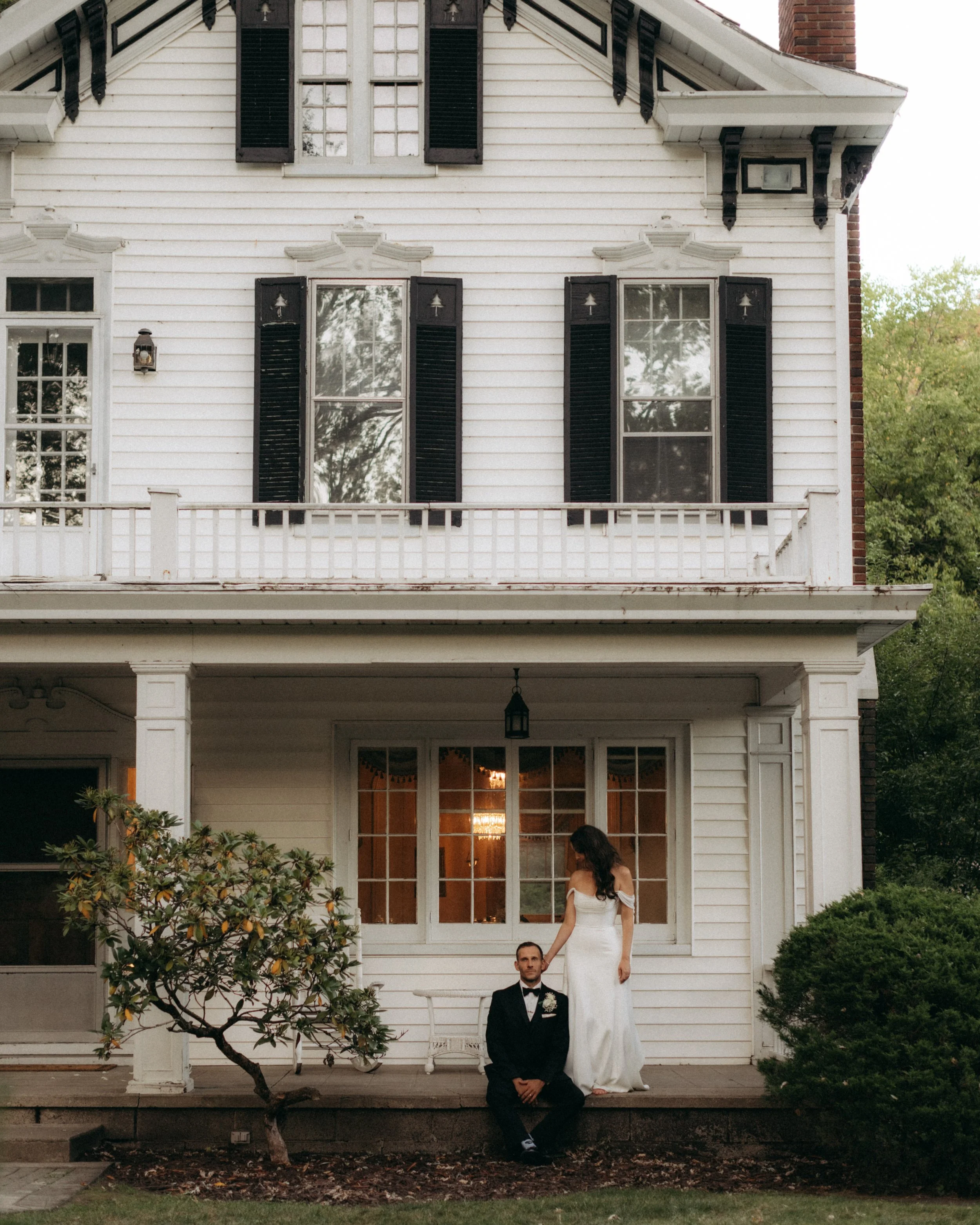 A newly married couple, woman in white wedding dress and man in black tuxedo, standing on the porch of a white house with black shutters. The woman is standing on a step above the man, who is seated, and they are holding hands.