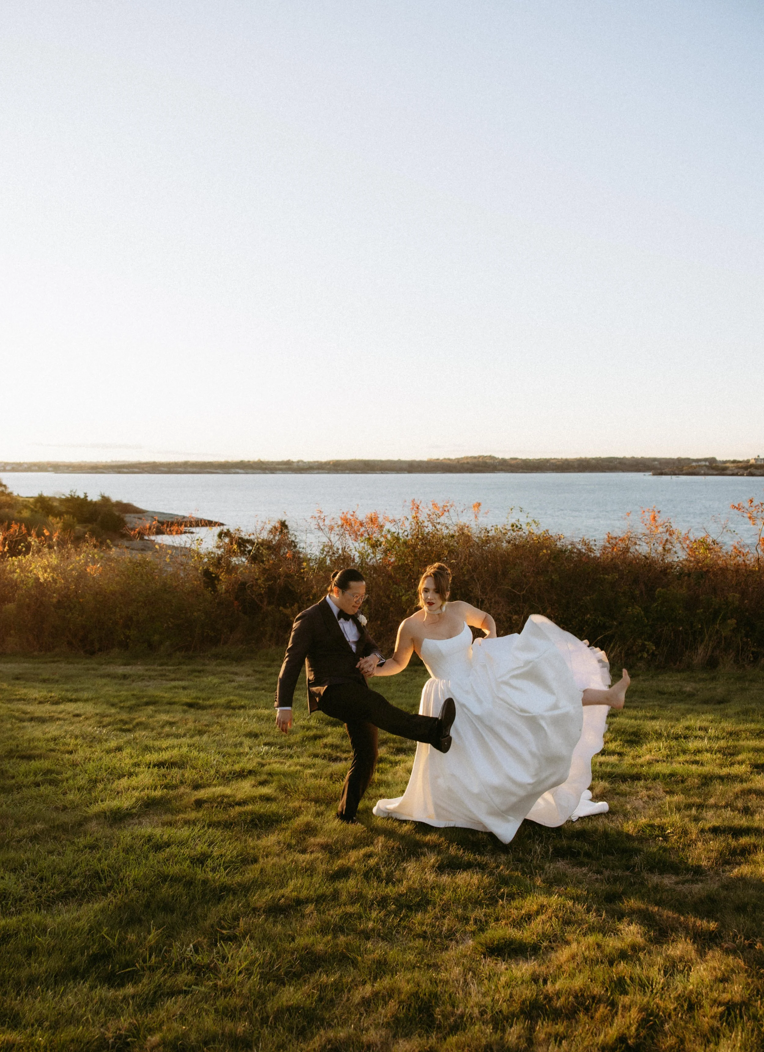 A bride and groom dancing outdoors at sunset near a body of water with greenery and trees in the background.
