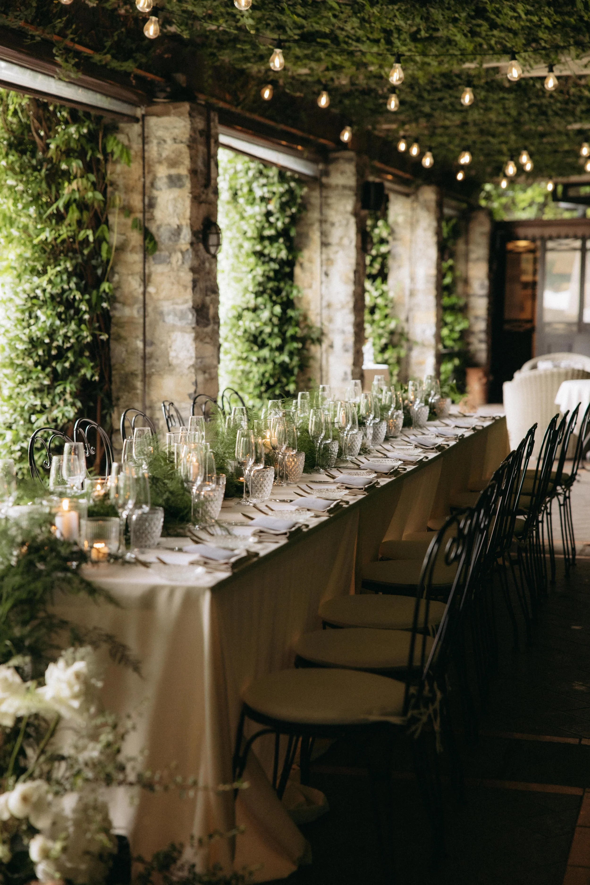 A long banquet table set for an event, decorated with greenery and lit candles, situated under a ceiling with string lights, in a rustic setting with exposed brick columns and greenery climbing the walls.