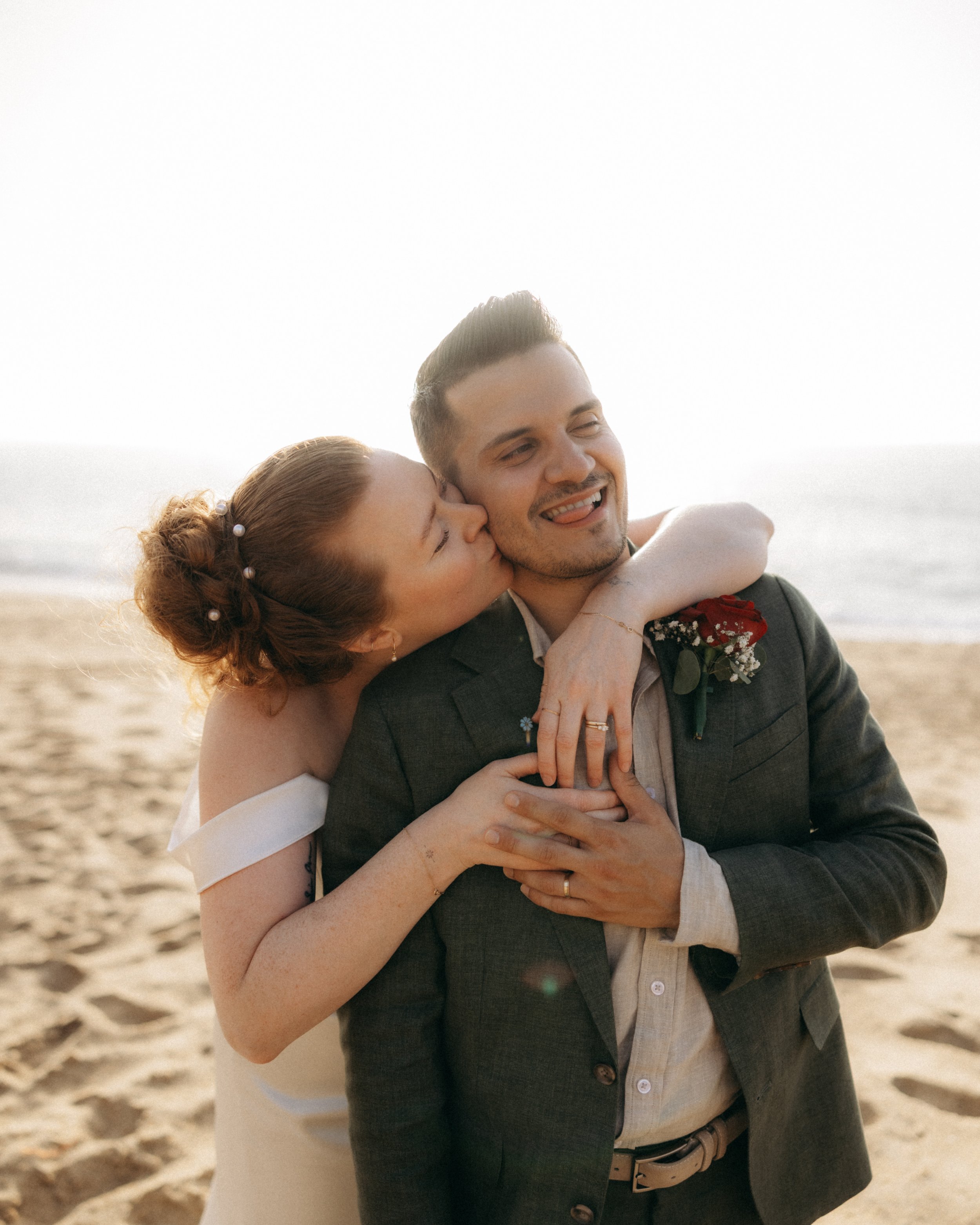 A couple on a beach embracing, with the woman kissing the man's cheek as he smiles with his tongue out.