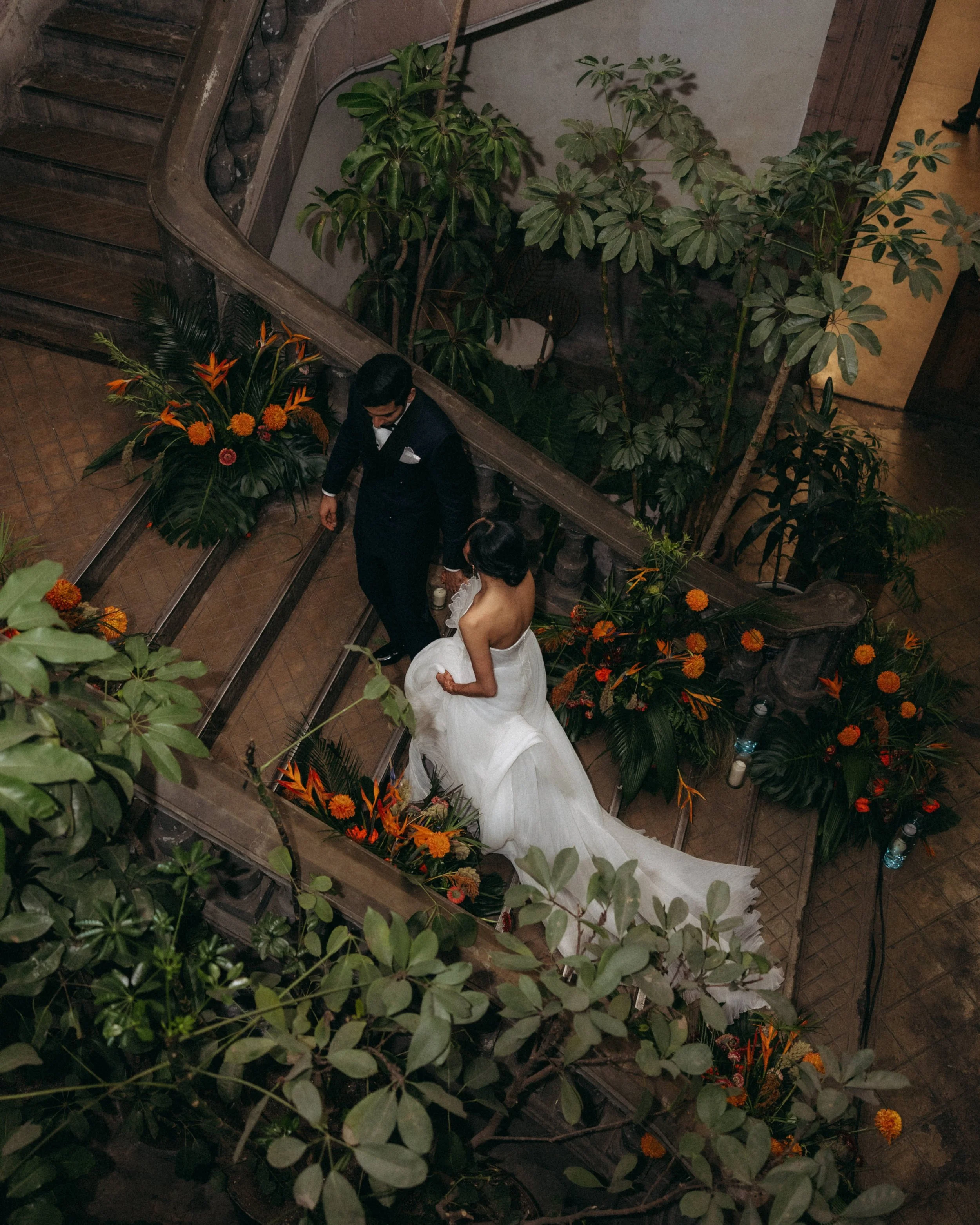 A bride and groom descending a staircase decorated with orange flowers, surrounded by lush green plants, during a wedding celebration.