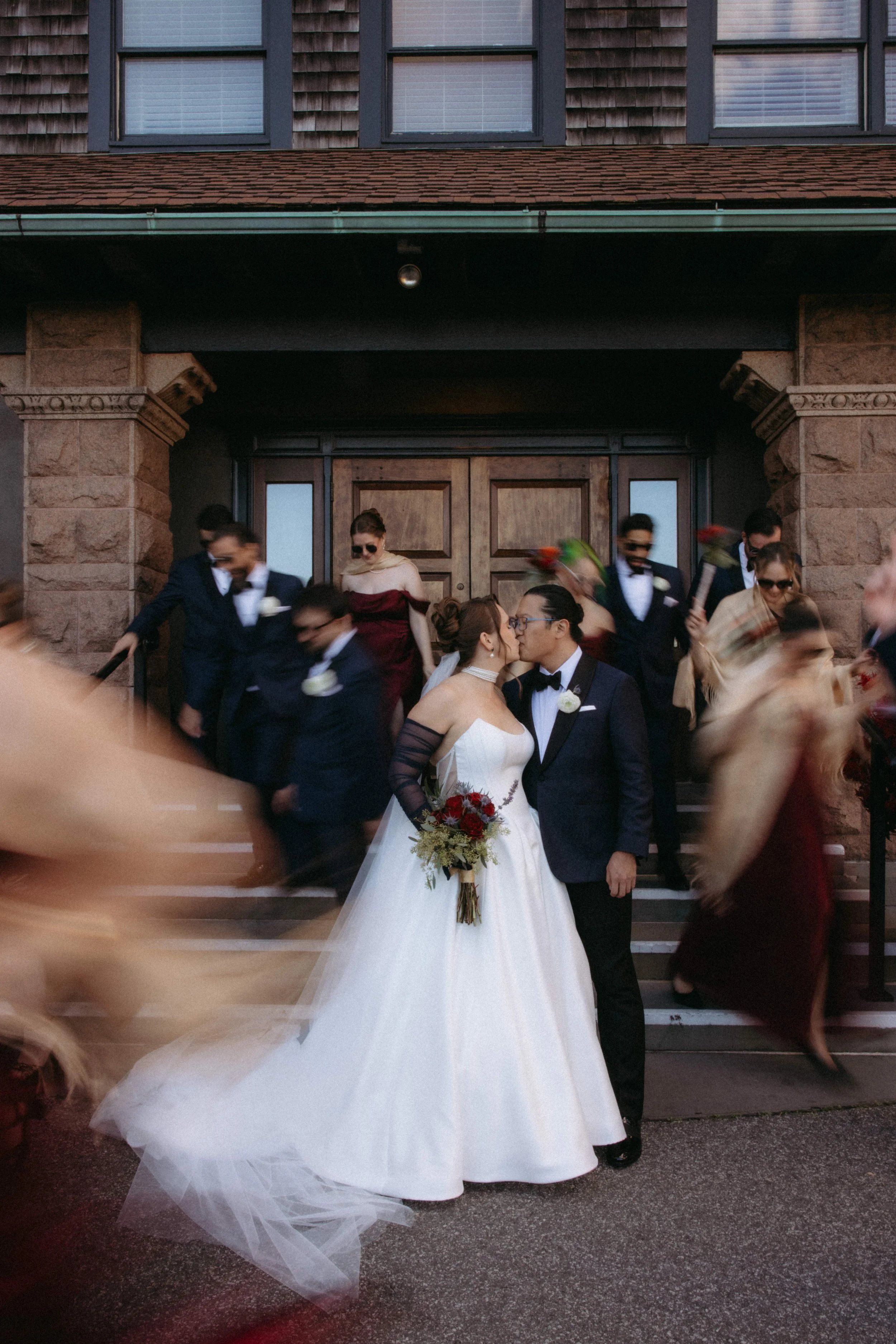 A bride and groom kissing in front of a church, surrounded by guests. The bride is wearing a white wedding gown and holding a bouquet of red and green flowers; the groom is dressed in a black tuxedo with a bow tie.