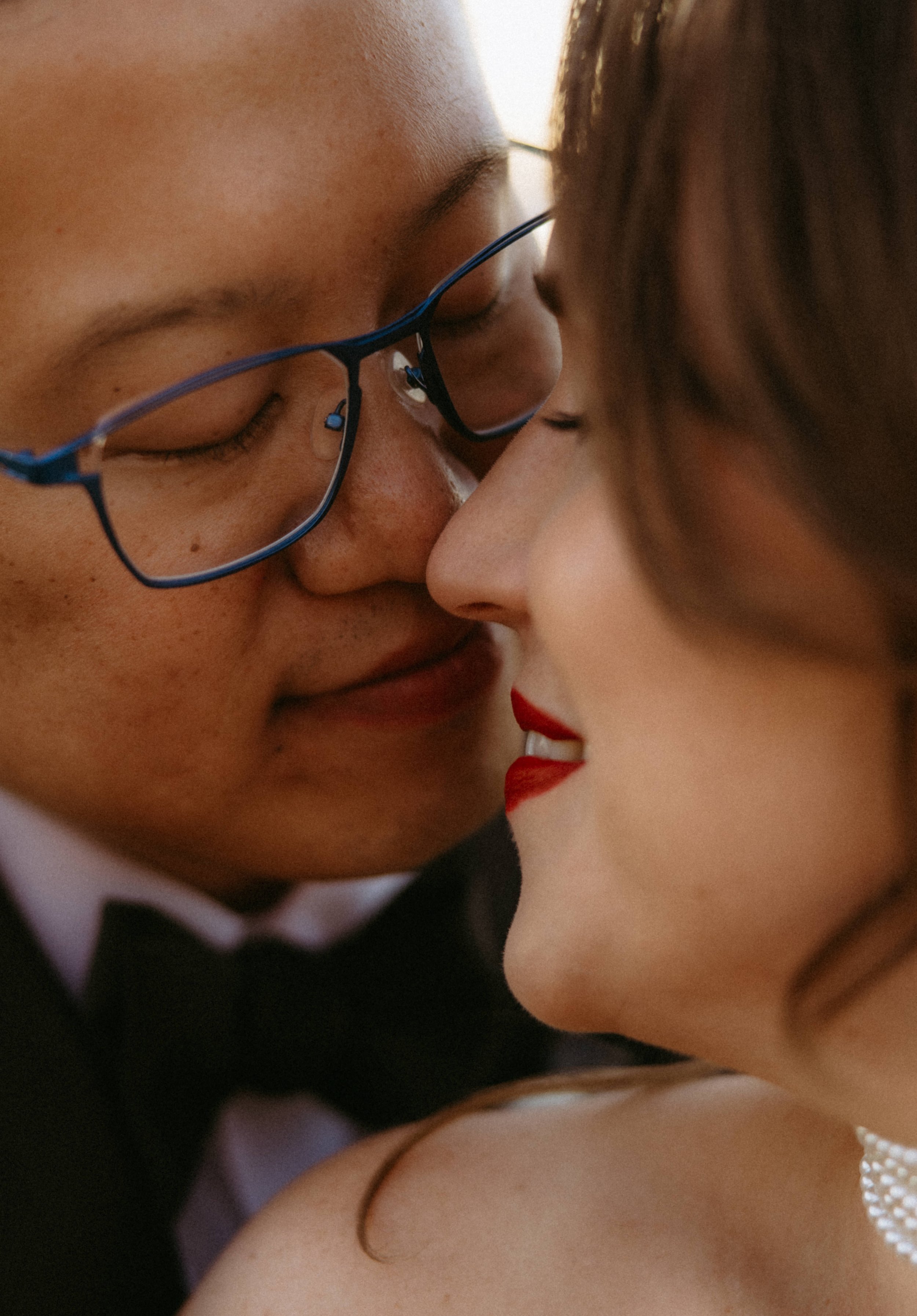 Close-up of two women touching foreheads, one wearing glasses and the other with bright lipstick, in an intimate moment.