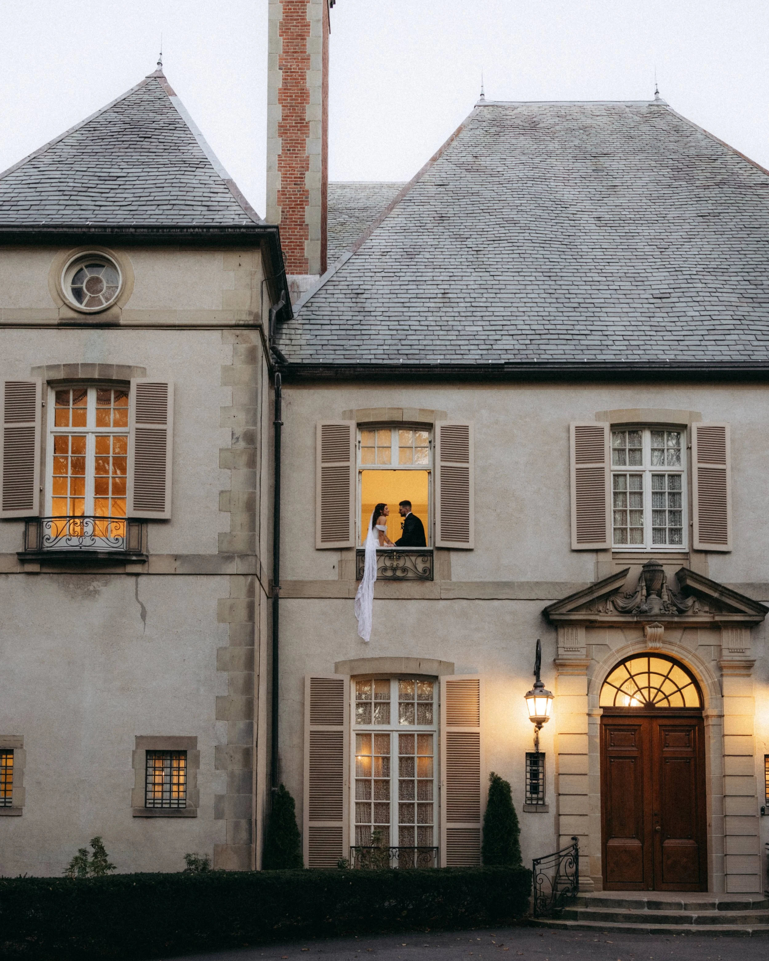 A couple is standing in front of an elegant residential building at dusk. The building features multiple tall windows with wooden shutters, a sloped slate roof, a brick chimney, and a rounded arched entrance with a wooden door. The couple, a bride an