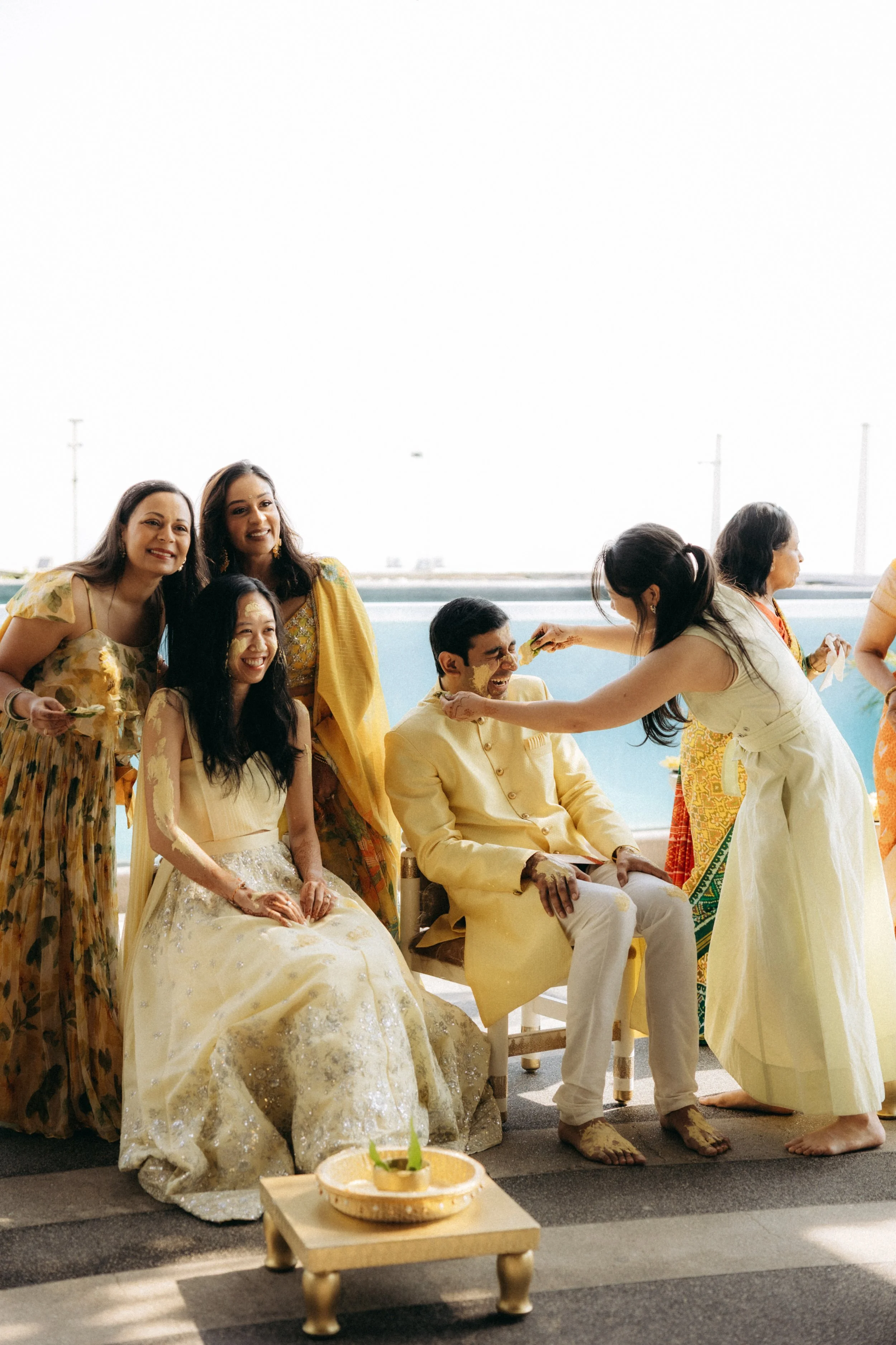A group of people participating in a traditional Indian wedding ceremony, with a bride in a yellow bridal gown sitting and a groom in cream attire sitting beside her, surrounded by friends and family dressed in colorful traditional clothing, outdoors