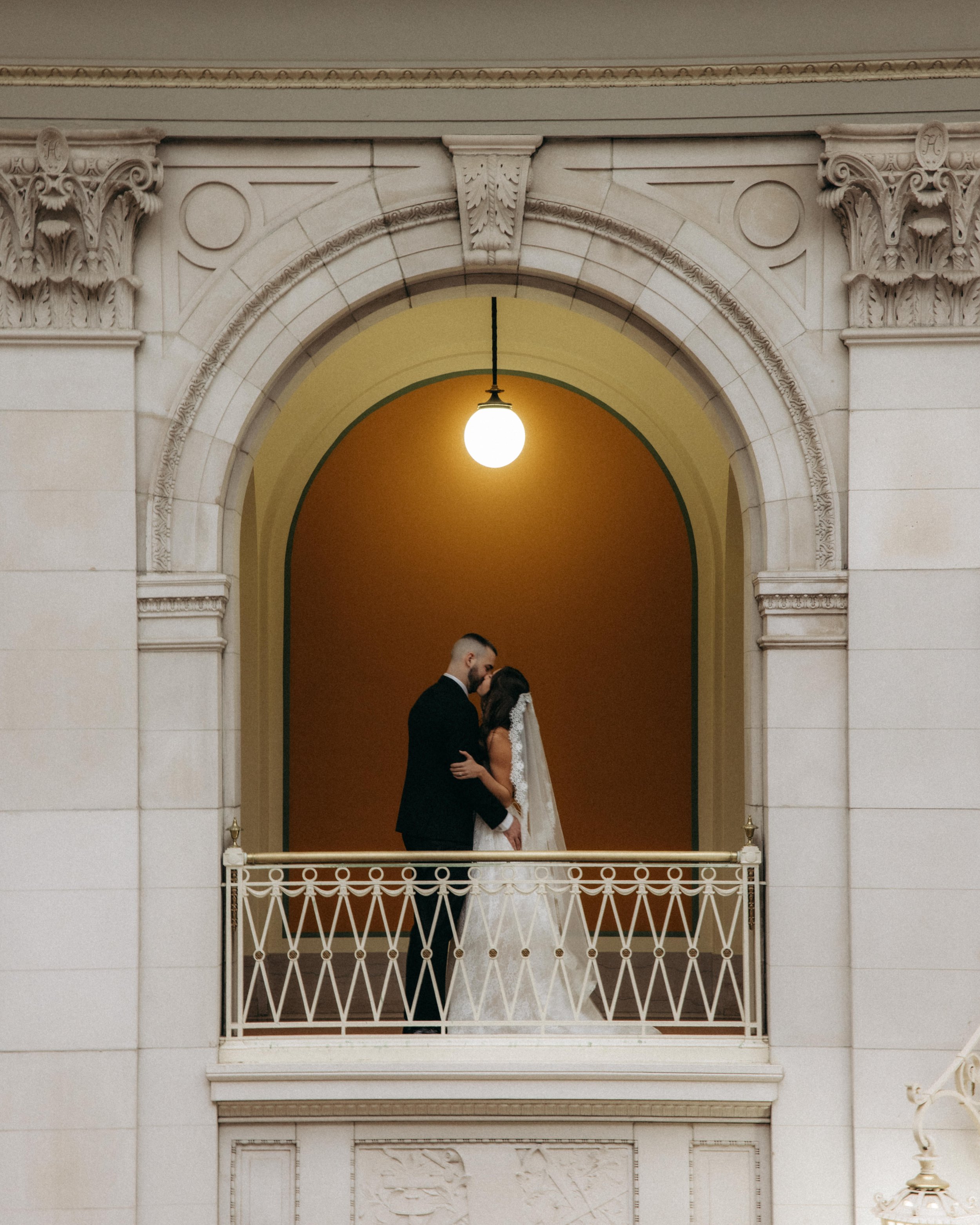 A couple in formal wedding attire sharing a kiss on a balcony inside a building with ornate architectural details, a large hanging light, and a warm interior background.