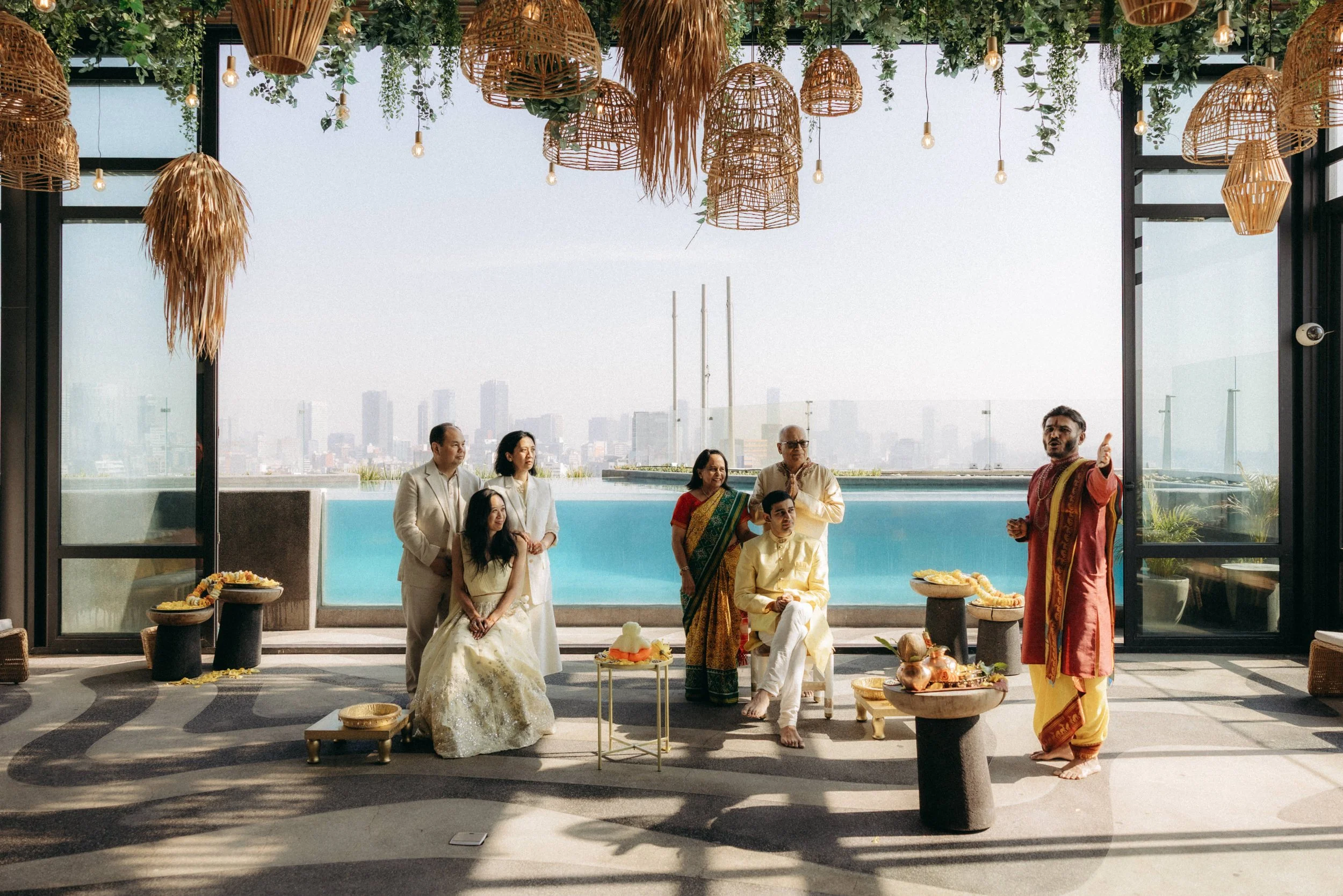 A group of people dressed in traditional Indian clothing participating in a cultural or religious ceremony indoors, with decorative hanging wicker lamps and greenery above them, and cityscape visible through a large window in the background.
