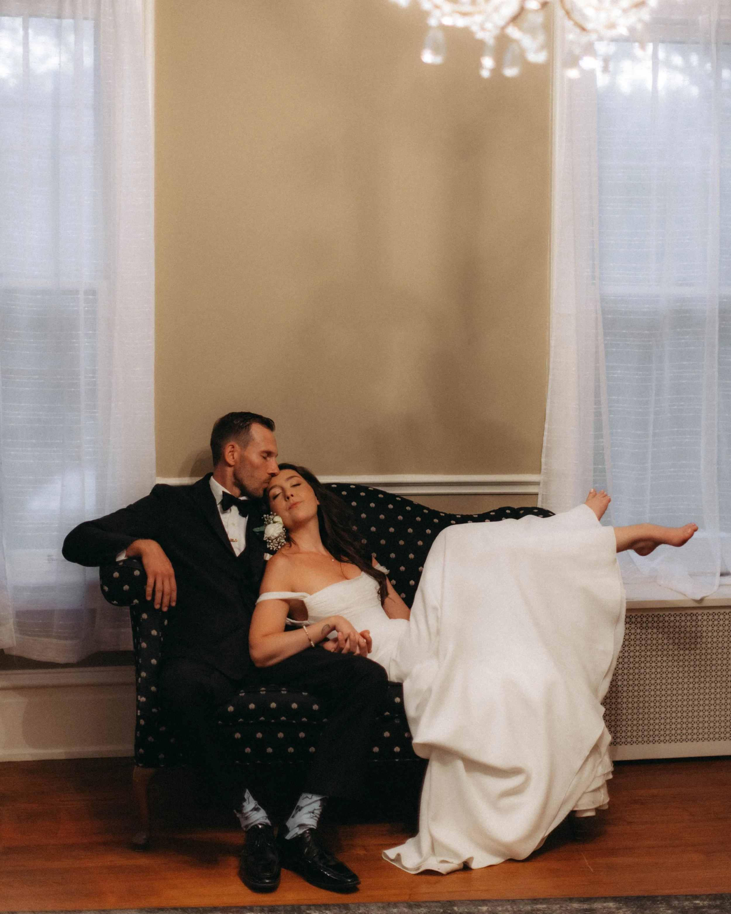 A couple dressed in formal attire, the man in a tuxedo and the woman in a wedding dress, are sitting on a black polka dot sofa in a well-lit room with sheer white curtains and a chandelier. The woman is lying with her eyes closed, and the man is lean