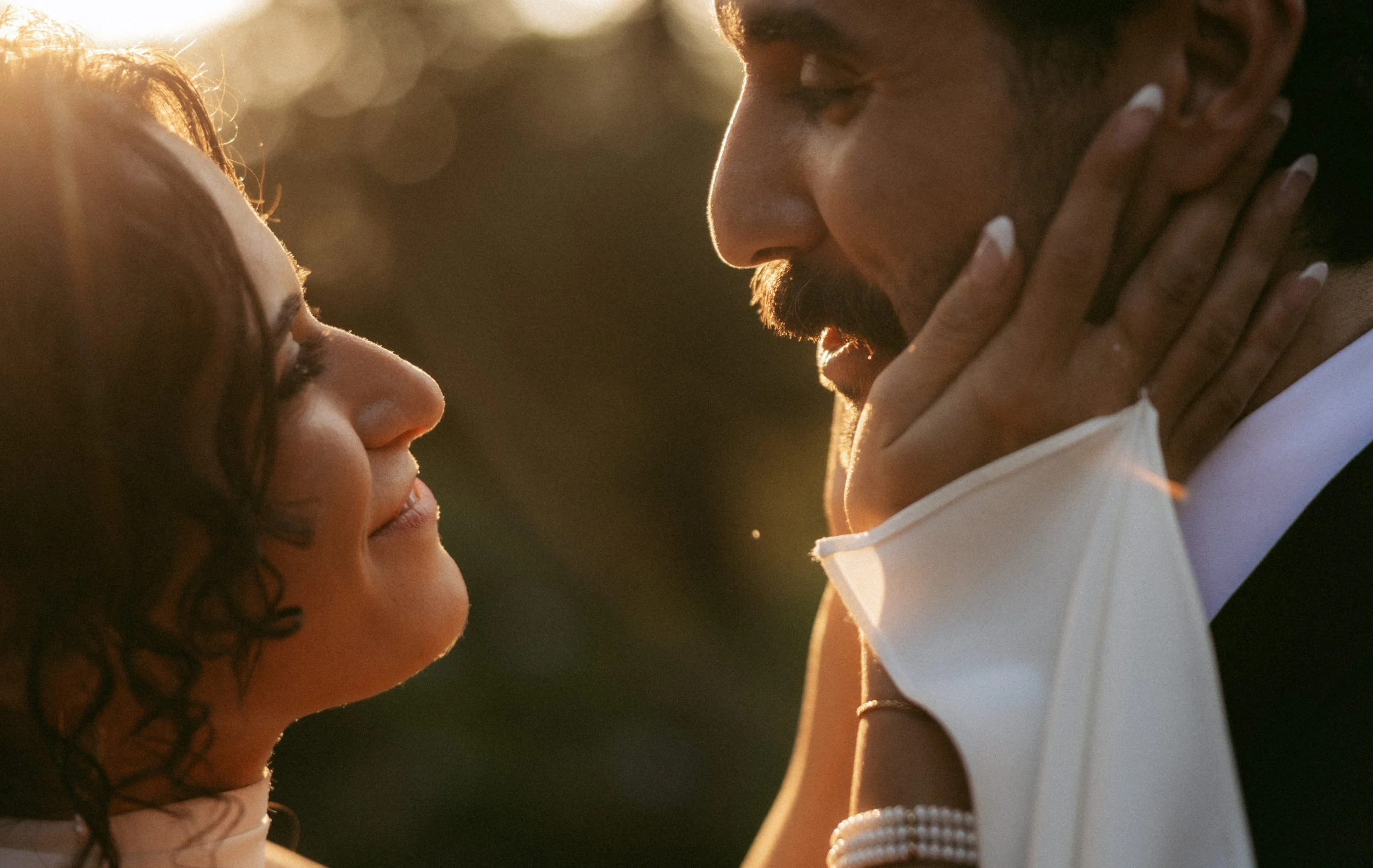 A man and a woman are close to each other outdoors during sunset, gently touching foreheads, sharing an intimate moment, with warm sunlight illuminating their faces.