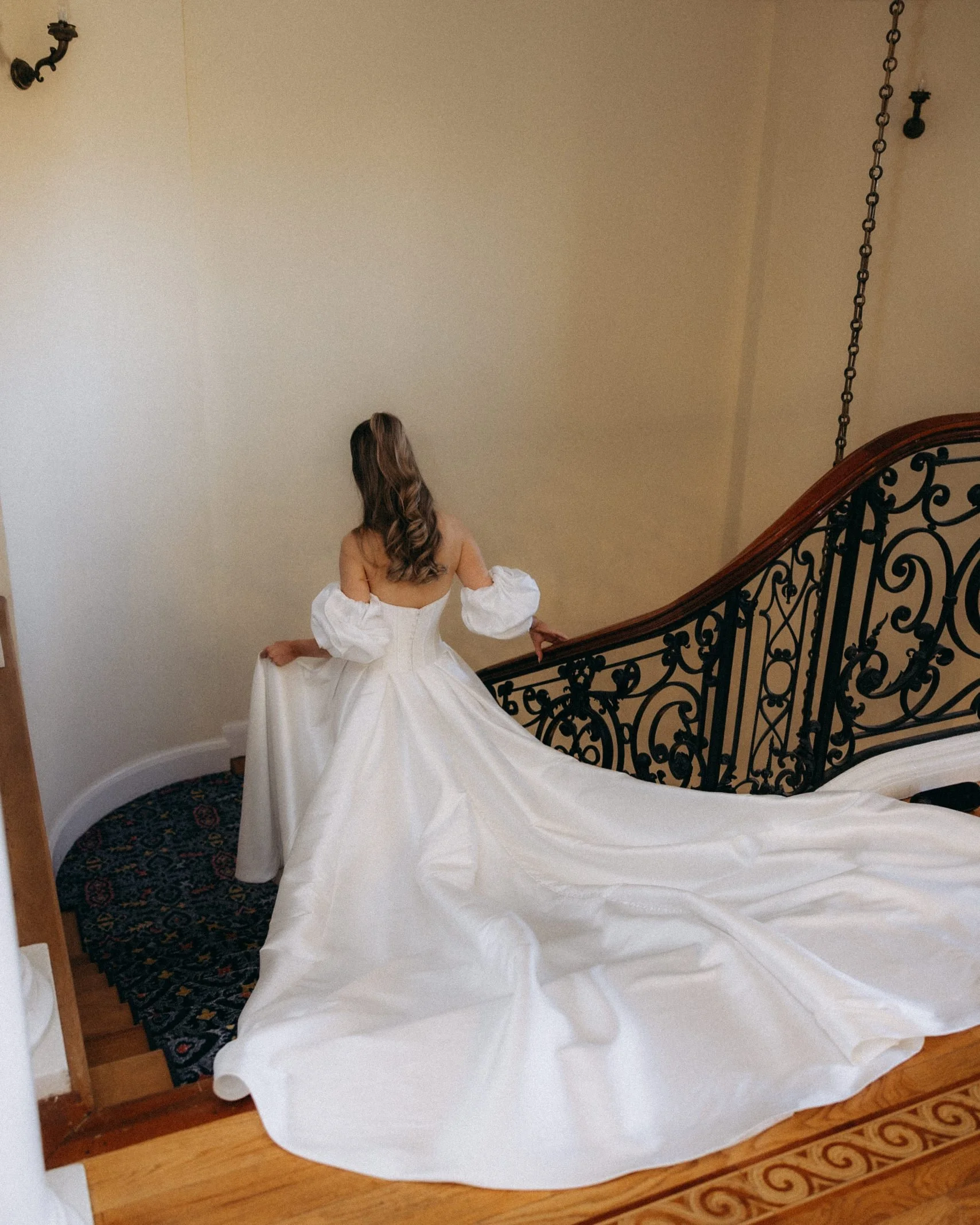 A woman in a white wedding dress sitting on a staircase with a black ornate railing, facing away from the camera, with long curled hair.