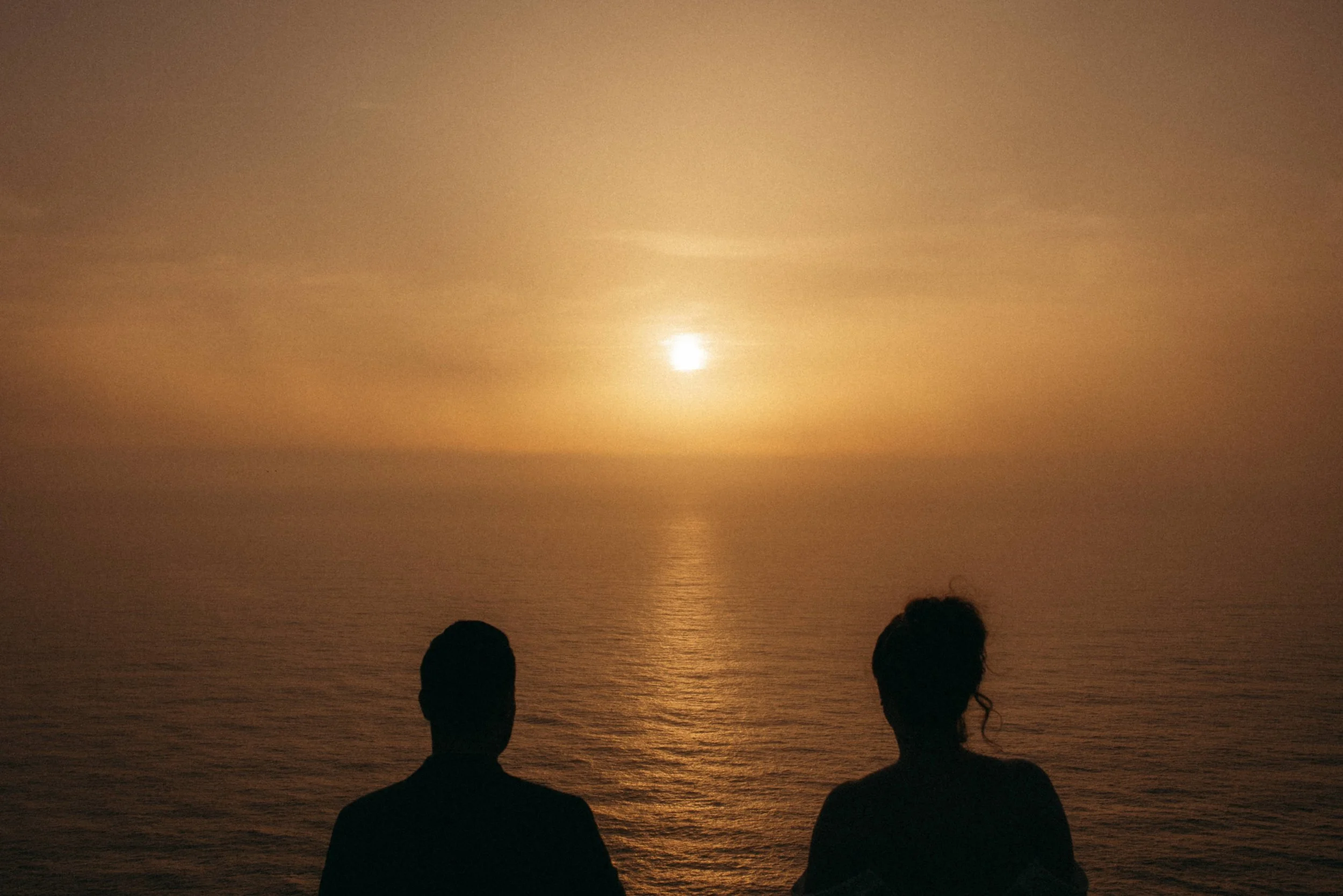 Two people sitting on the beach during sunset, facing the water with the sun low in the sky, creating a warm glow on the sea surface.