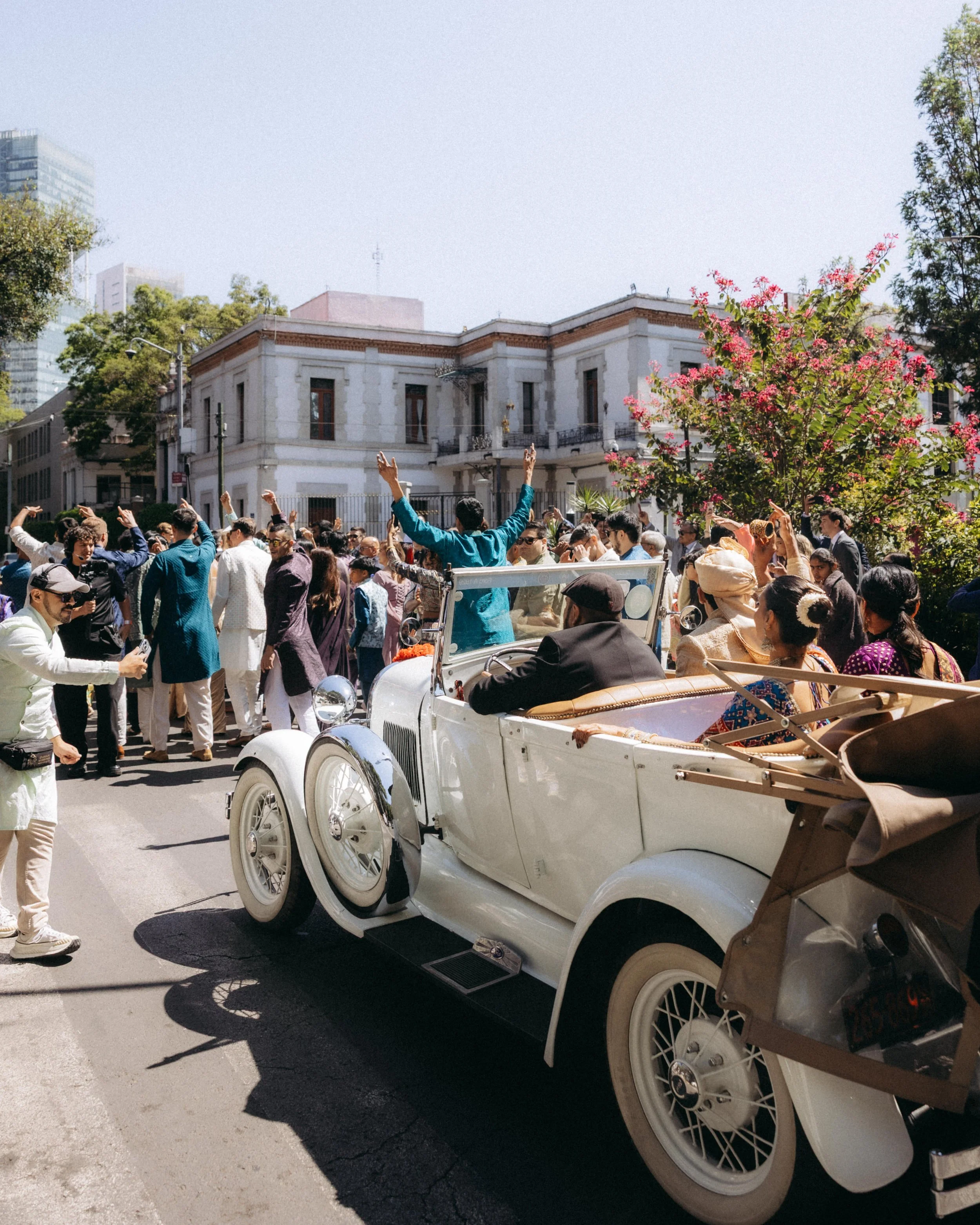 A crowd of people gathered on a city street, with some dancing and celebrating. There is a vintage white car in the foreground, and a historic building in the background, along with some tall modern buildings and green trees.