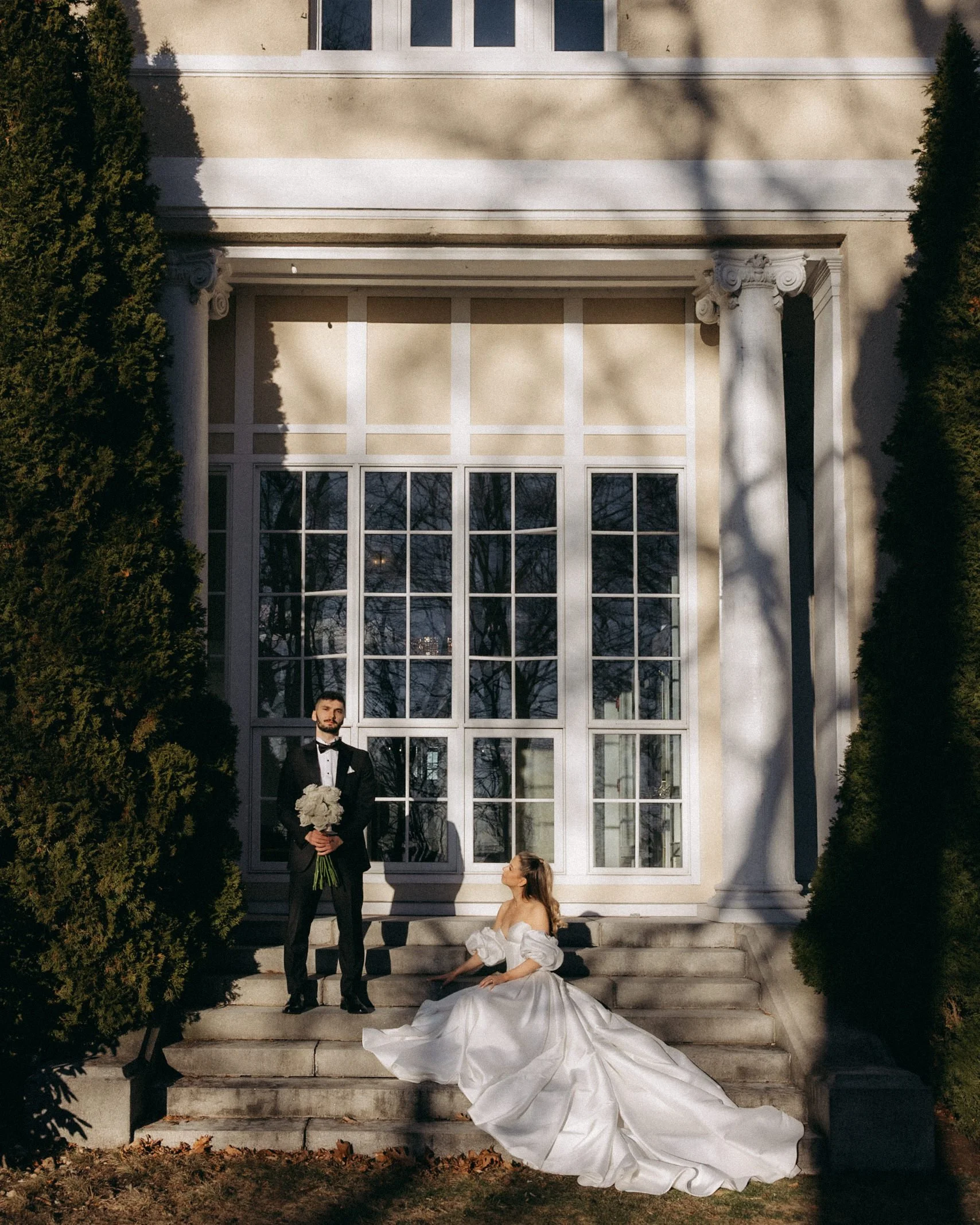 A bride sitting on the steps of a large house with a man in a tuxedo standing beside her, holding a bouquet of white flowers, with a woman in a wedding dress kneeling in front of him, outside on a sunny day.
