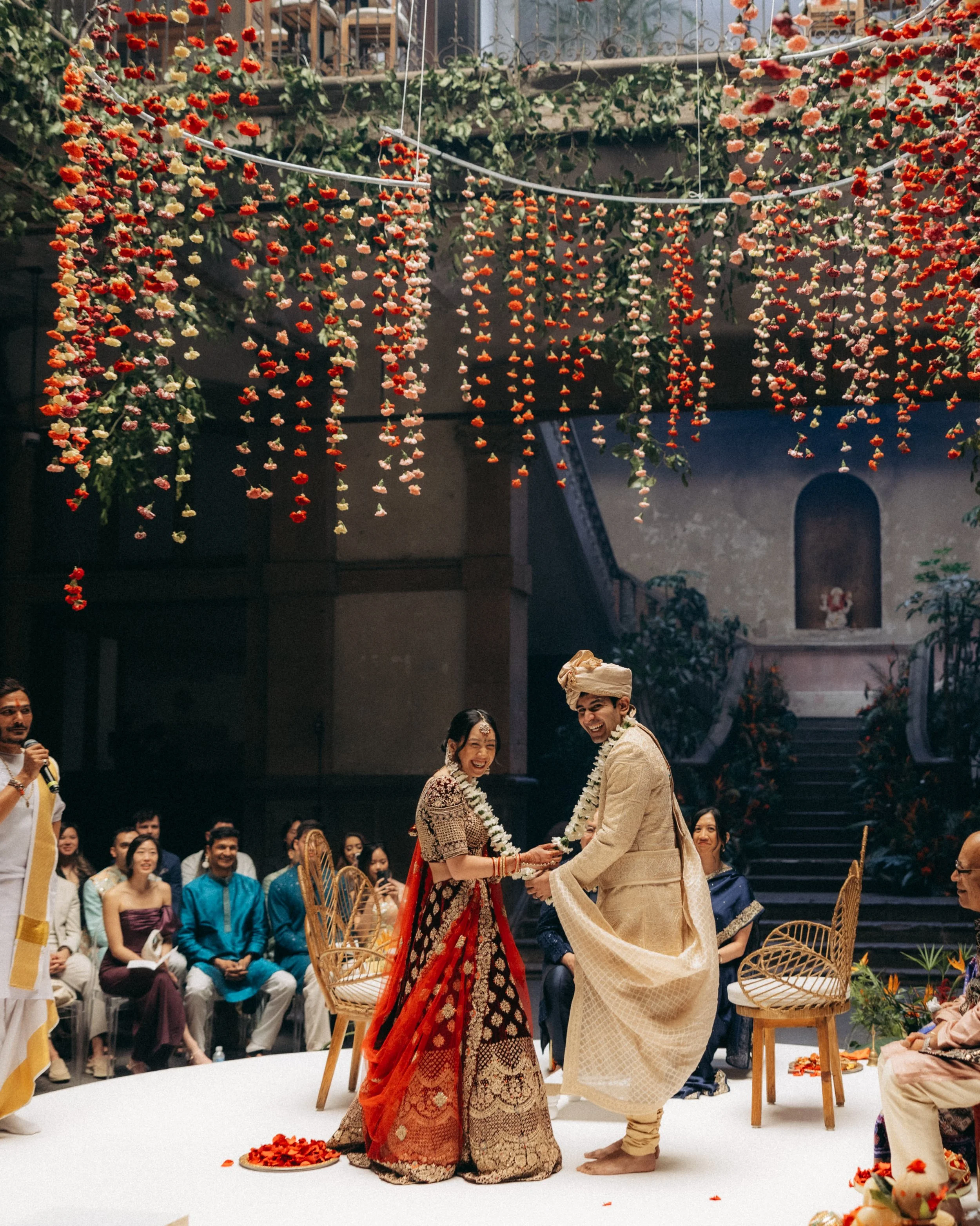 A traditional Indian wedding ceremony with the bride and groom exchanging garlands, surrounded by guests. The bride is in a red and gold lehenga, and the groom is in cream-colored traditional attire. The setting is decorated with hanging flowers and 