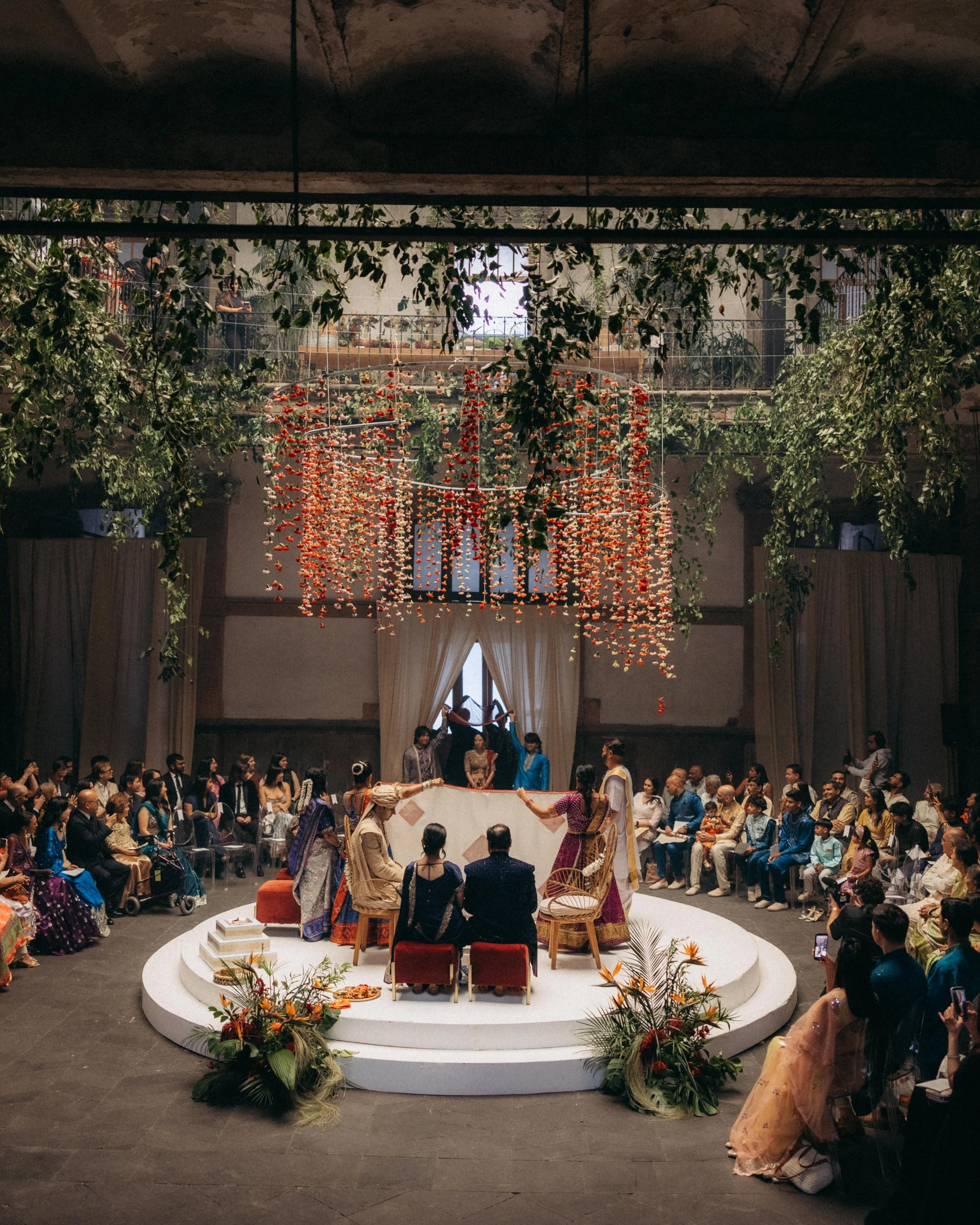 People attending a traditional Indian ceremony or celebration indoors, with a woman in a saree and women in colorful dresses around a raised platform, floral decorations, and hanging floral arrangements.