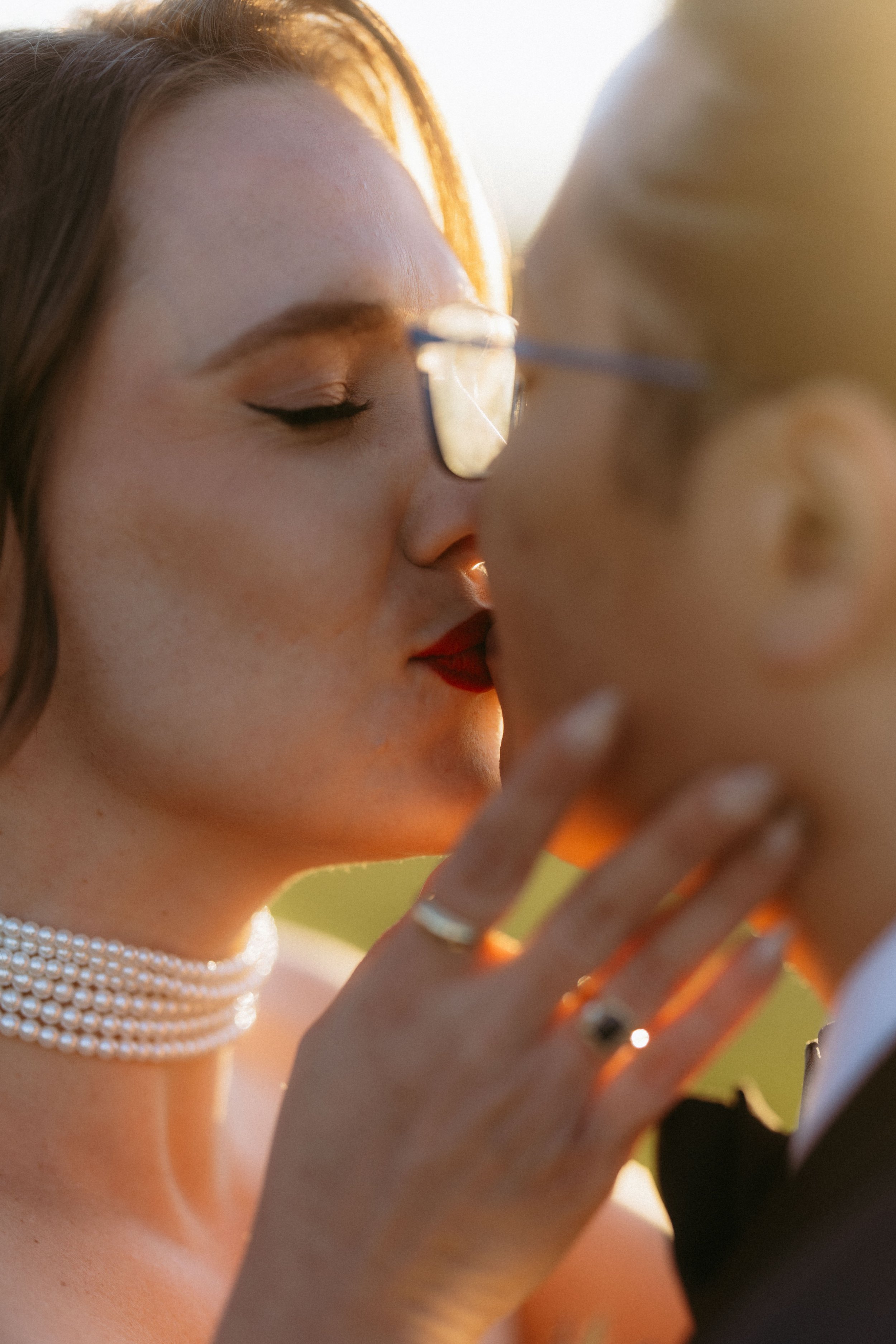 Close-up of a woman with red hair, closed eyes, and makeup, holding a mirror with her hand. She is wearing a pearl necklace and rings on her fingers. The background is blurred with warm lighting.
