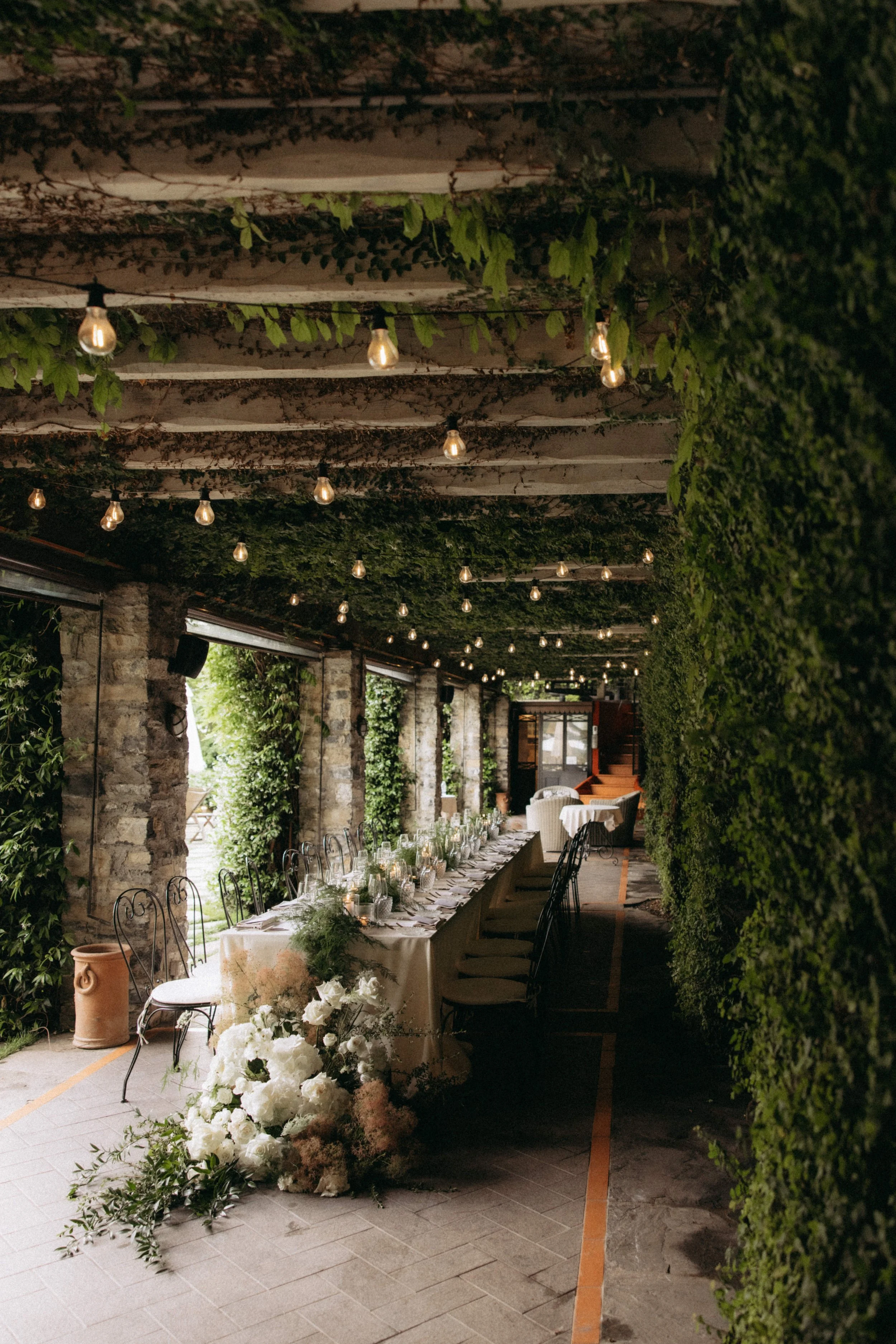 Elegant outdoor event space with a long table set for a formal dinner, decorated with floral arrangements and candles. The area features stone walls, green foliage, string lights hanging from a wooden pergola, and large windows.