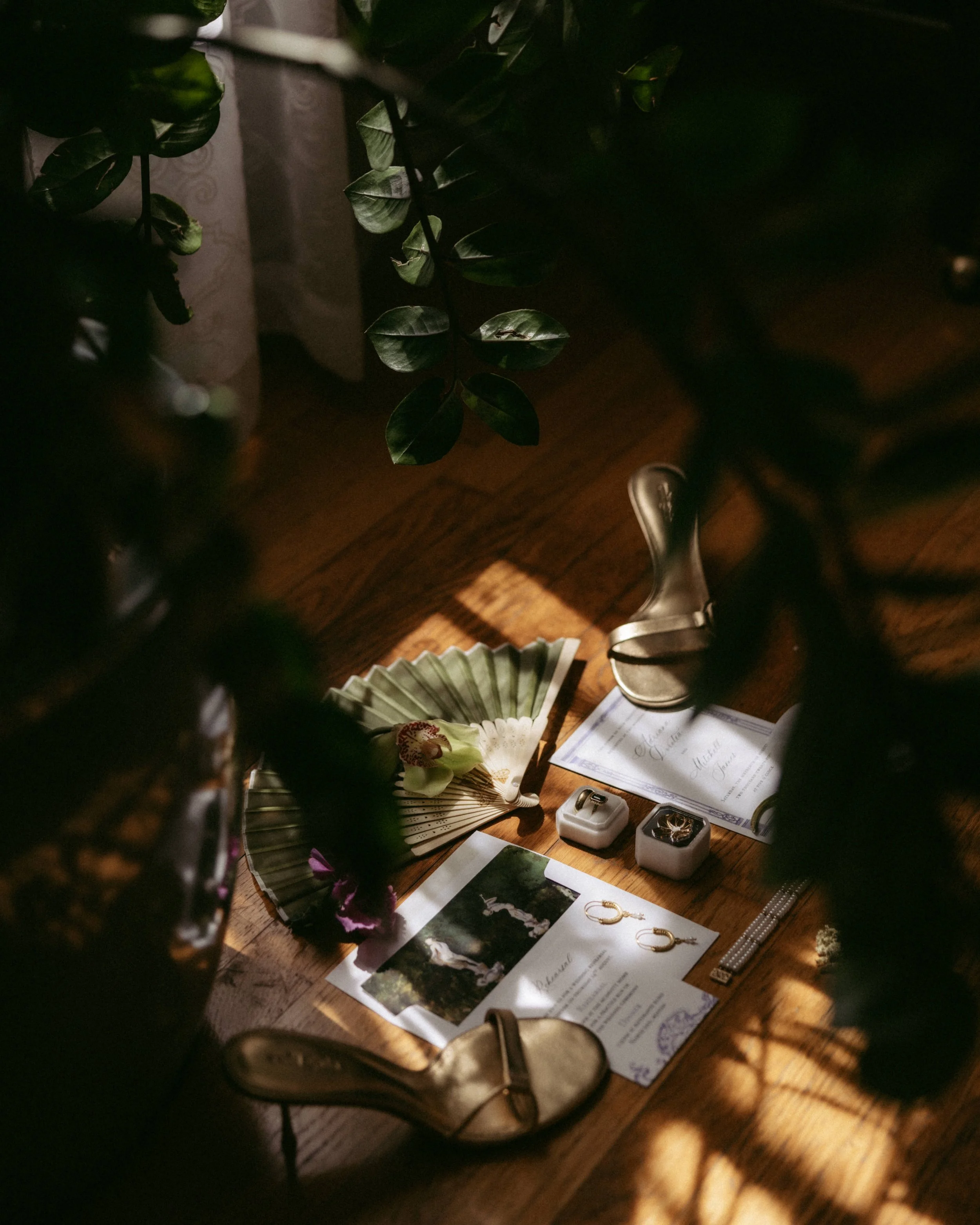 A wooden table with scattered jewelry, printed papers, and a decorative paper fan. The jewelry includes rings, earrings, and watches. There are also some plants and shadows visible on the table.