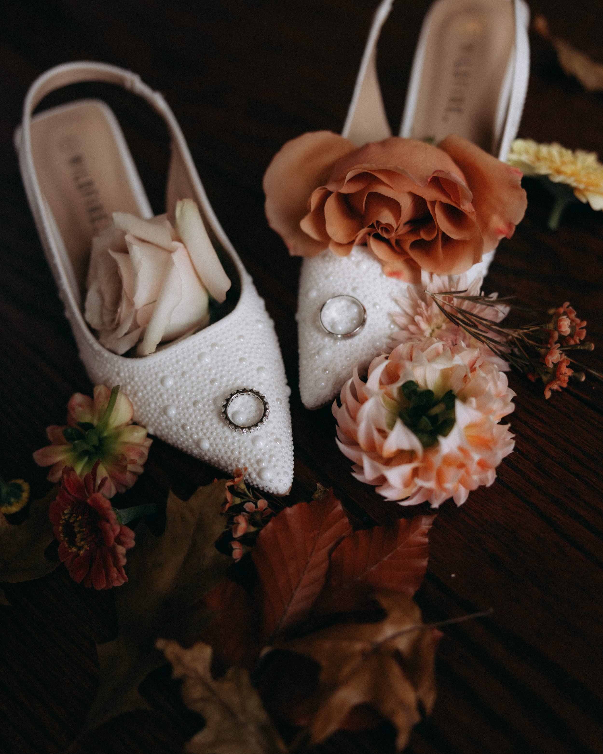 A pair of white high-heeled shoes with peep toes, decorated with pearl embellishments and two rings placed on the toes. Surrounding the shoes are orange and pink flowers and brown autumn leaves, set on a dark wooden surface.