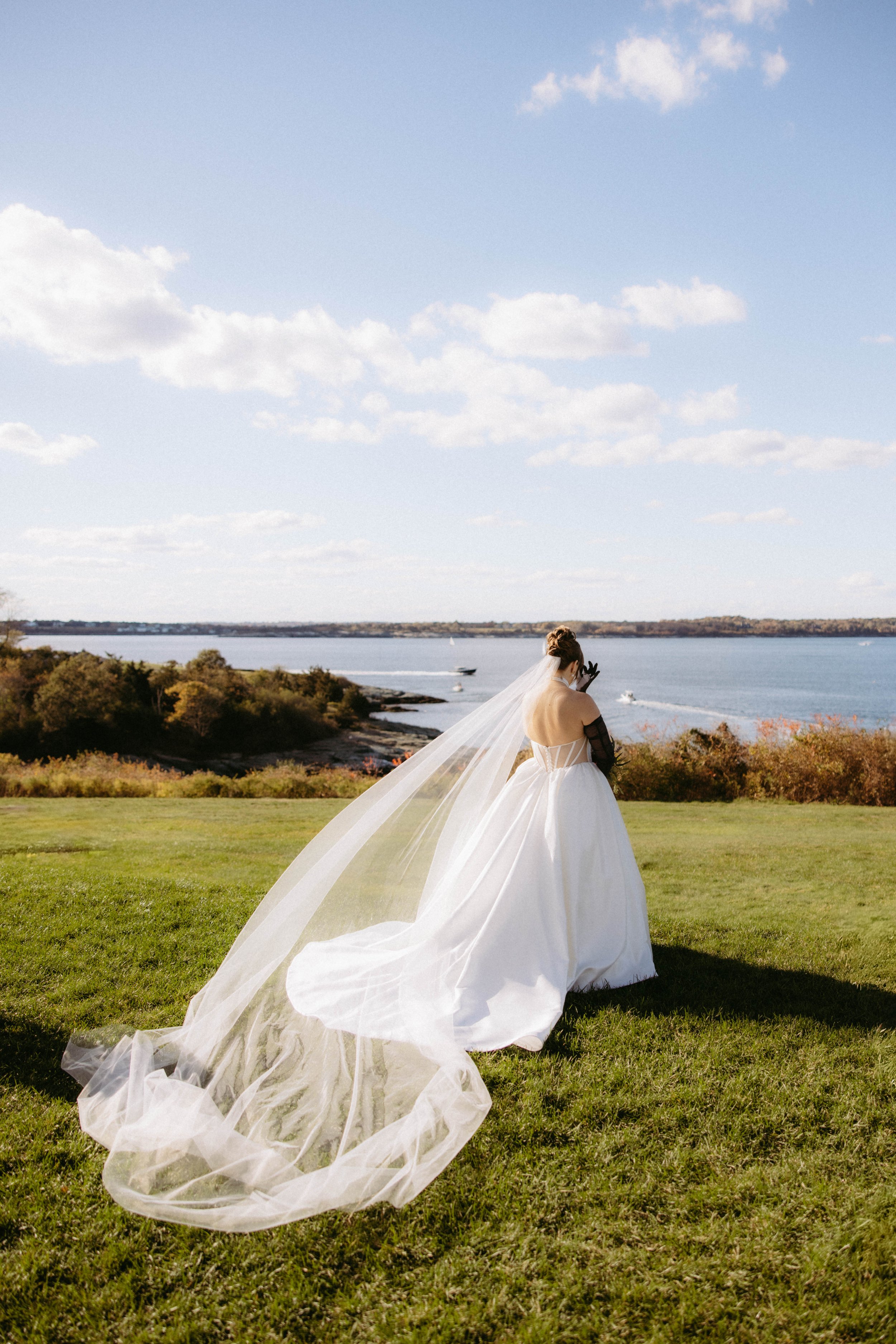 A bride in a white wedding gown and veil standing on a grassy area near the water, with trees and boats in the background, under a blue sky with scattered clouds.