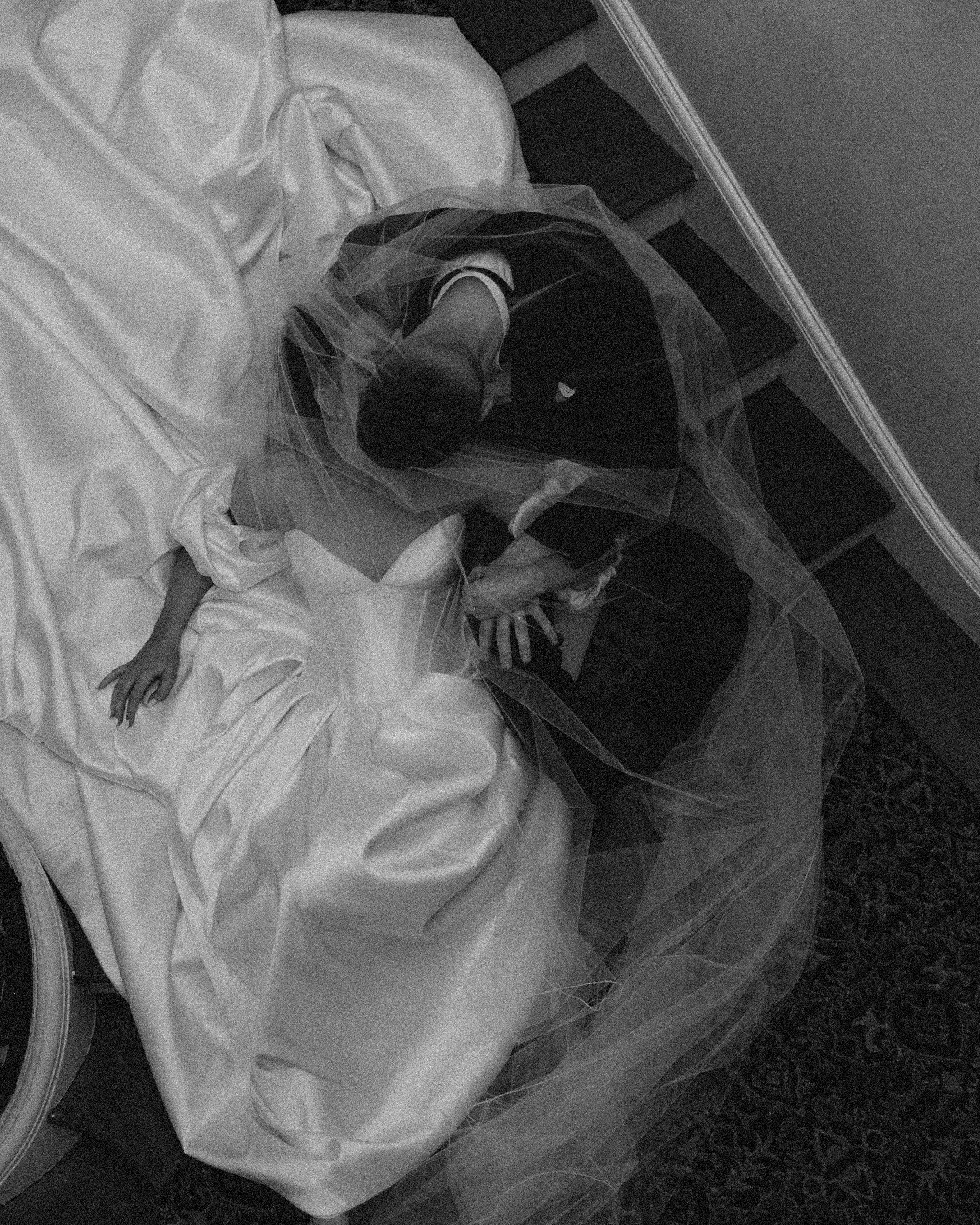 A person lying on a hospital bed with their leg elevated, wearing a black Adidas sneaker, wrapped in a protective plastic sheet, with a ceiling and wall visible in the background.