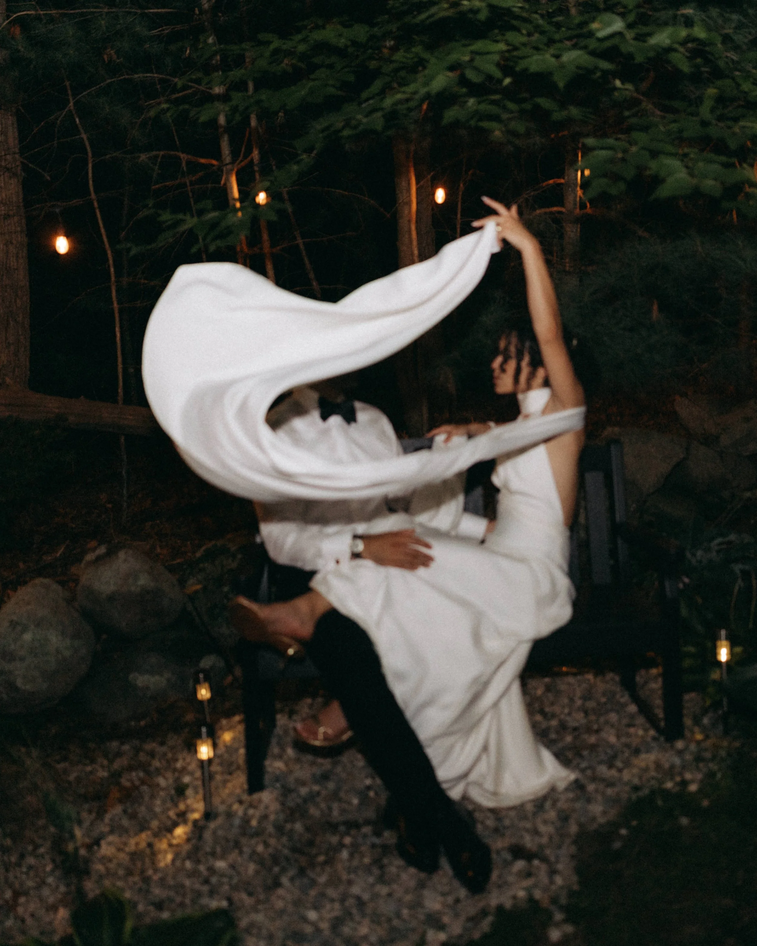 A person wearing a wedding dress and a tuxedo is dancing outdoors at night with a woman dressed in formal attire, surrounded by string lights and trees.
