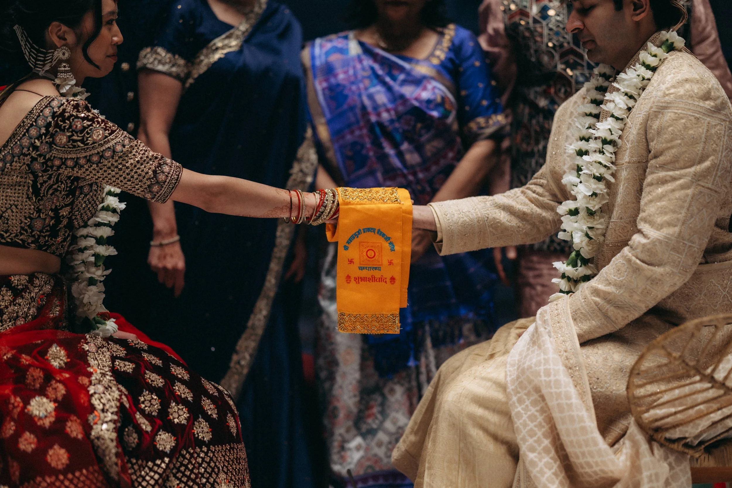 A traditional Indian wedding ceremony where a bride and groom are exchanging floral garlands. The bride is wearing a richly embroidered maroon and gold outfit with jewelry, and the groom is in a cream-colored sherwani with a floral garland. They are 