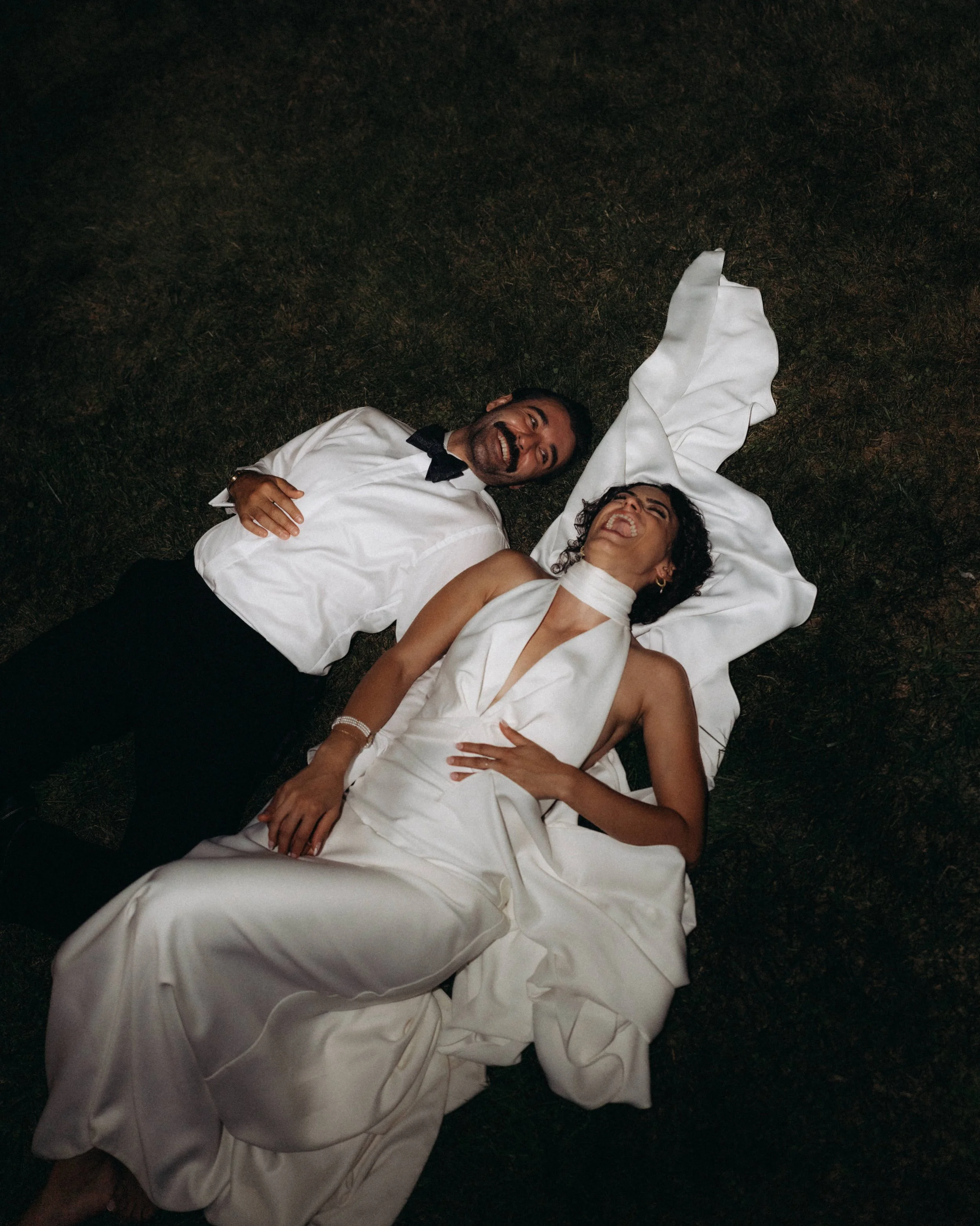 A man and a woman lying on the ground at night, smiling and laughing, dressed in formal white attire; the man is wearing a tuxedo and the woman is wearing a white dress with a slit.