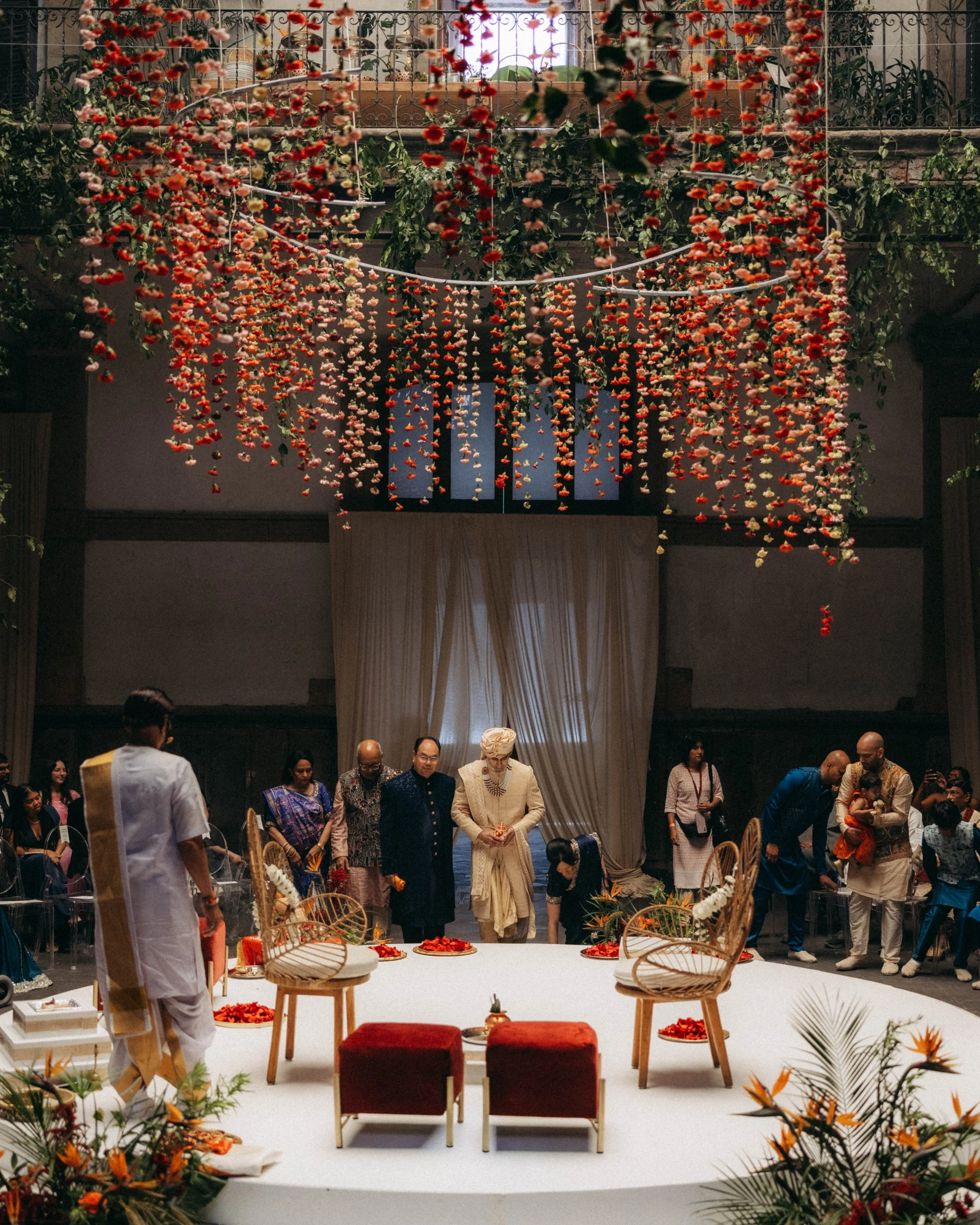 A wedding ceremony stage decorated with hanging red and pink flower garlands, with people dressed in traditional attire standing and sitting around the stage. Traditional chairs and cushions are placed on the stage, with decorative flowers around the