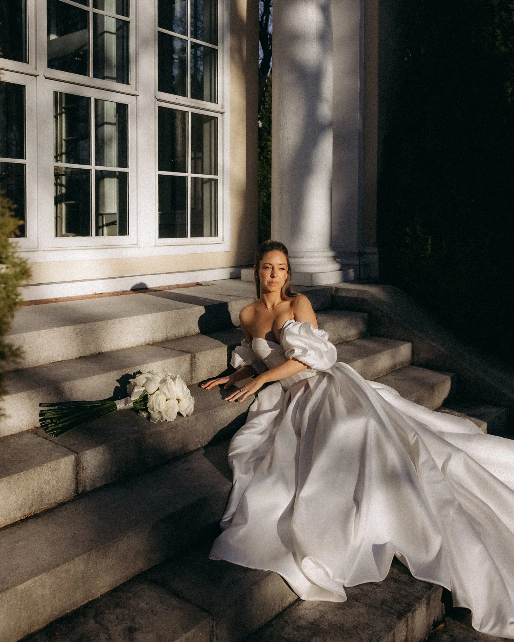 Woman in a white wedding dress sitting on steps outside a house with large white columns, sunlight illuminating her face and a bouquet of white roses beside her.