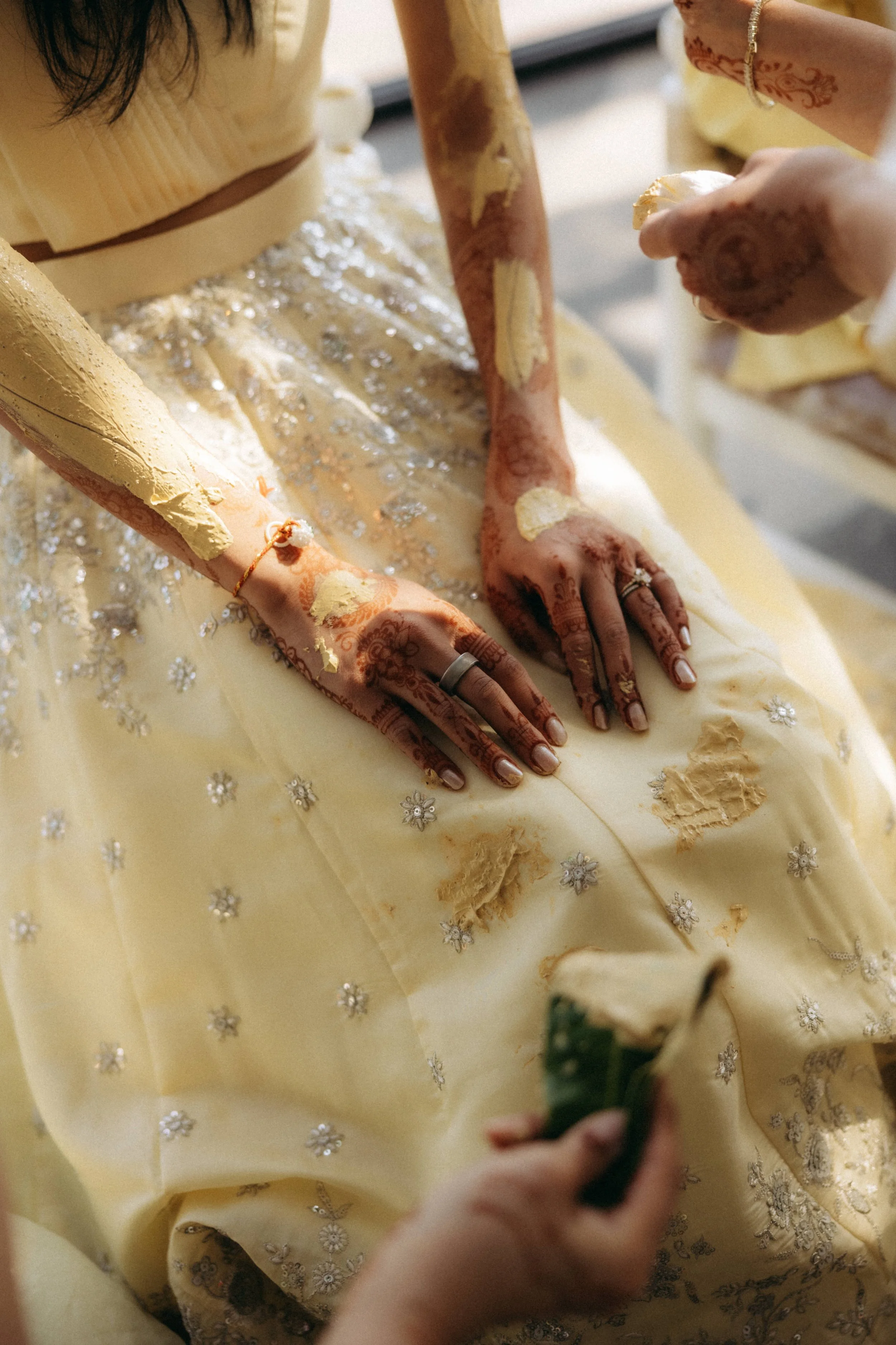 Close-up of a bride's hands with mehndi designs, wearing rings, and in a yellow dress with silver embroidery, while applying or touching her dress at a wedding.