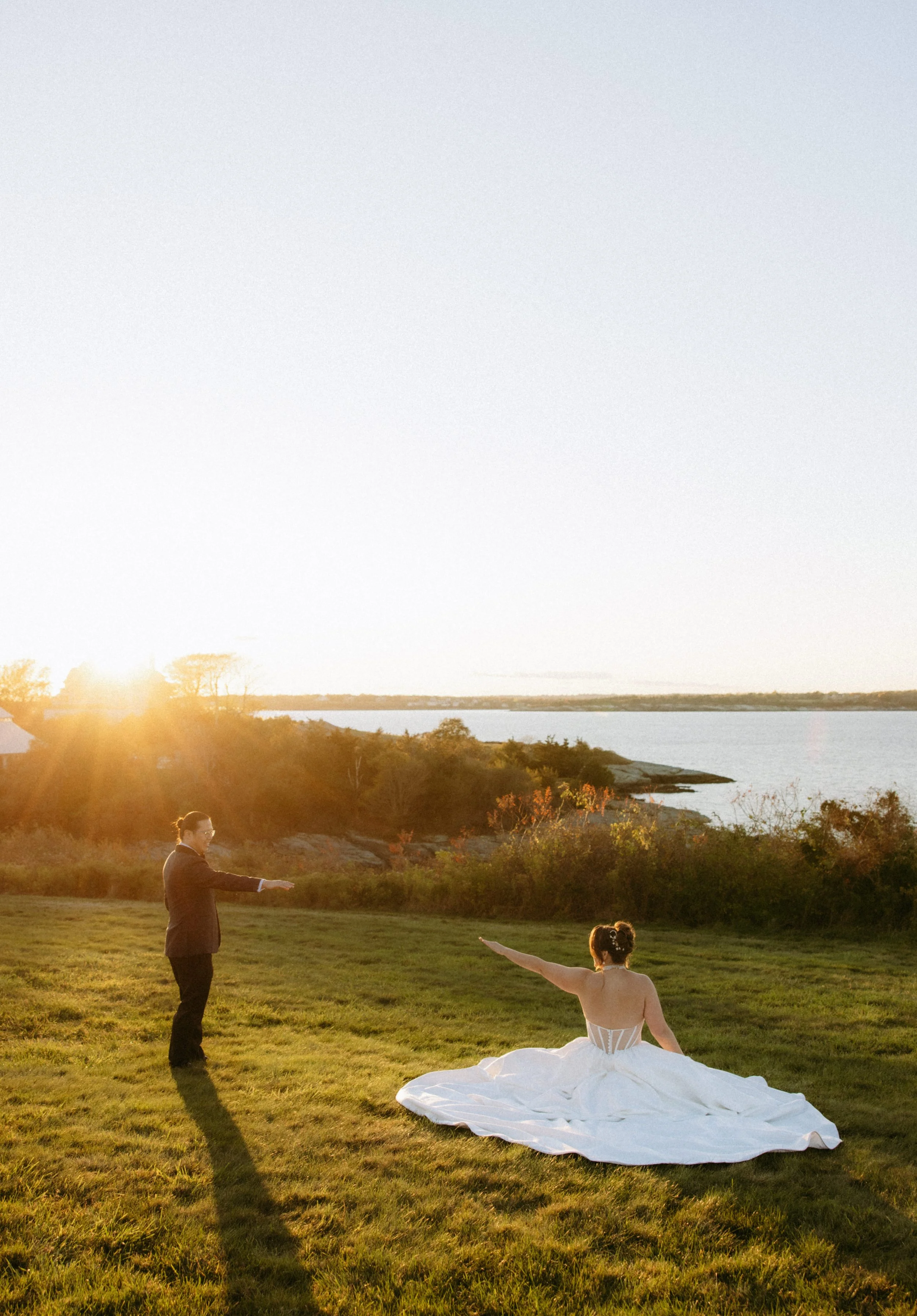 A wedding ceremony taking place outdoors on a grassy area near a body of water during sunset. A bride in a white wedding dress is seated with one arm raised, while a man in a dark suit stands nearby with his arm extended. Two children are present, bo