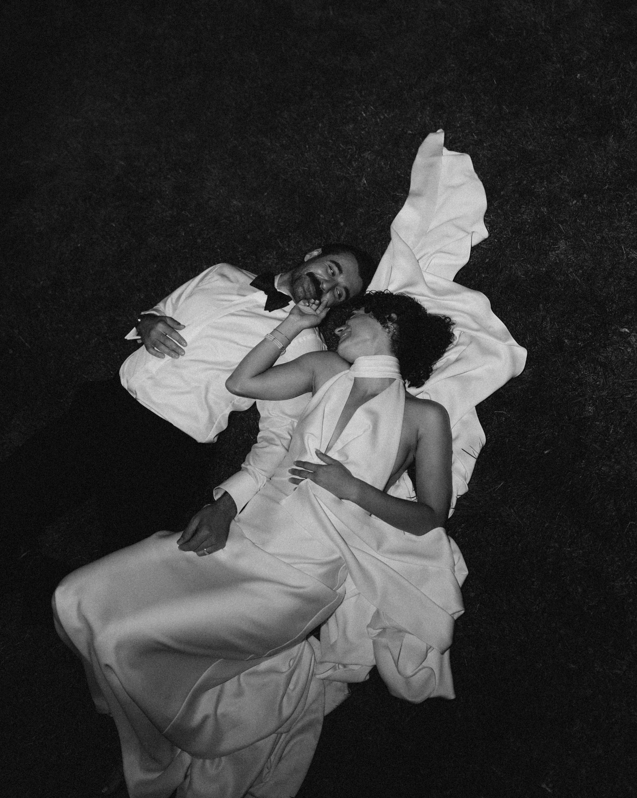 Black and white photo of a happy couple lying on the ground in wedding attire. The man is wearing a tuxedo with a bow tie, smiling and looking at the woman. The woman is in a wedding dress, resting her head on the man's shoulder, with her eyes closed