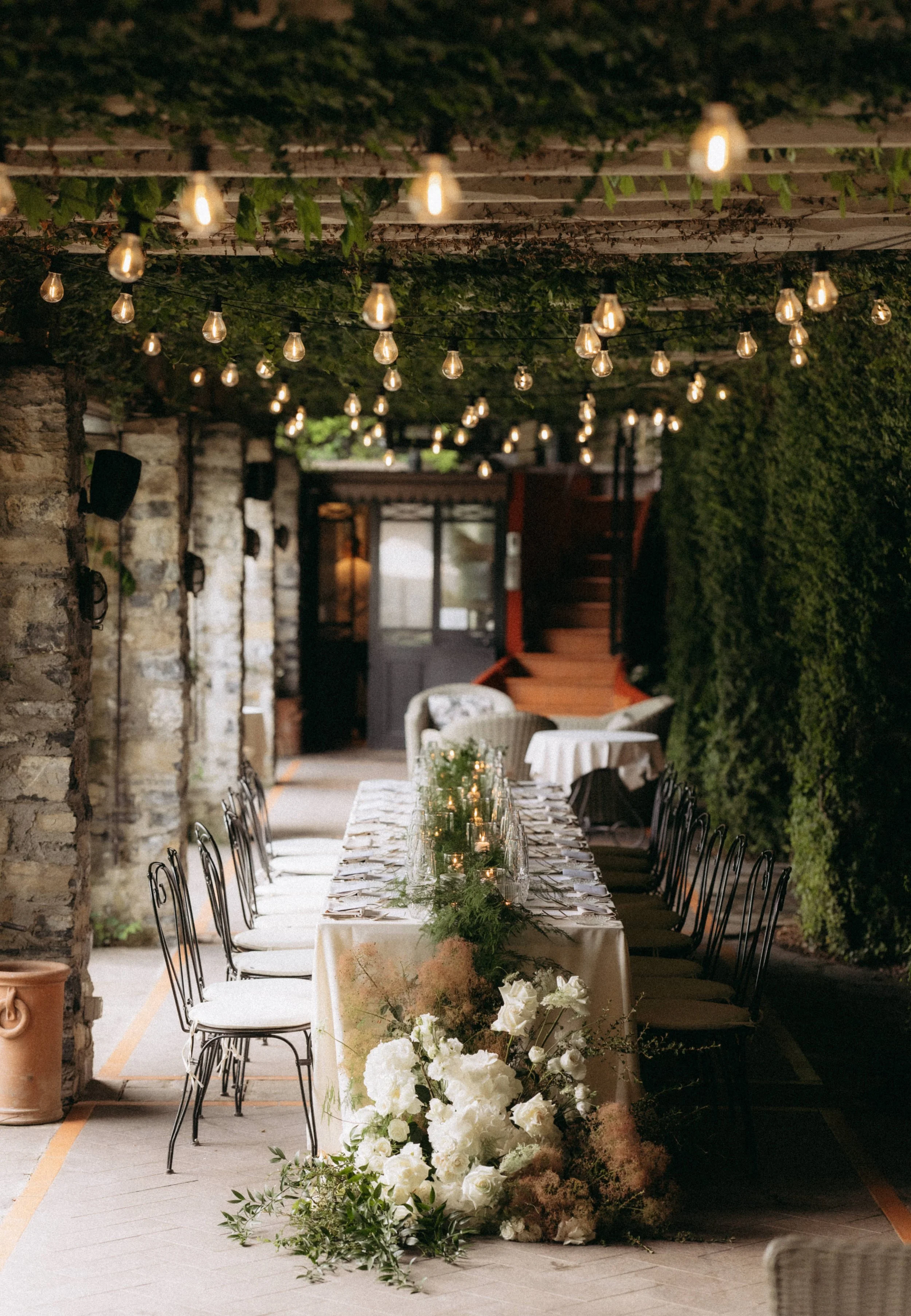 An outdoor dining area decorated for a special event with a long table set with white tablecloth, glassware, and candles in glass holders, surrounded by black chairs, with floral arrangements of white flowers and greenery. String lights hang overhead