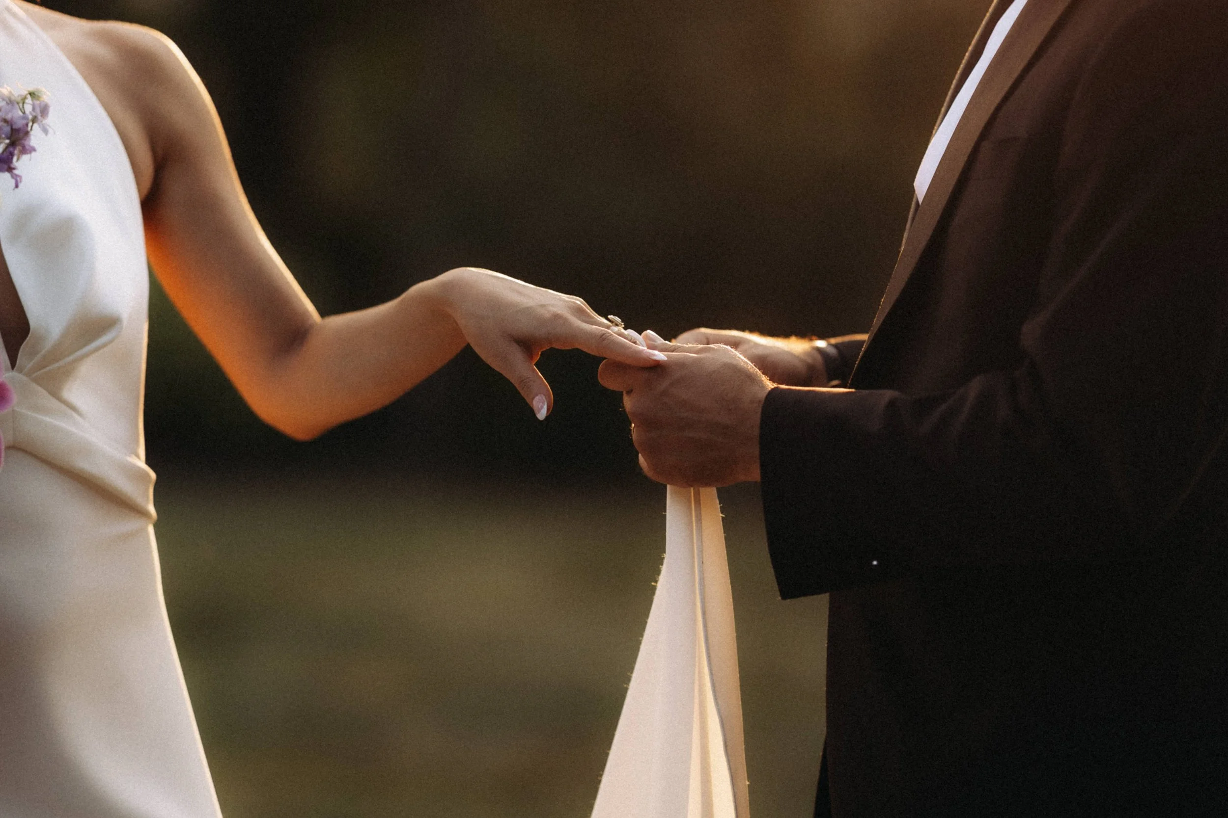 Close-up of a bride and groom holding hands during a wedding ceremony, with the bride wearing a white dress and the groom in a dark suit, in an outdoor setting at sunset.