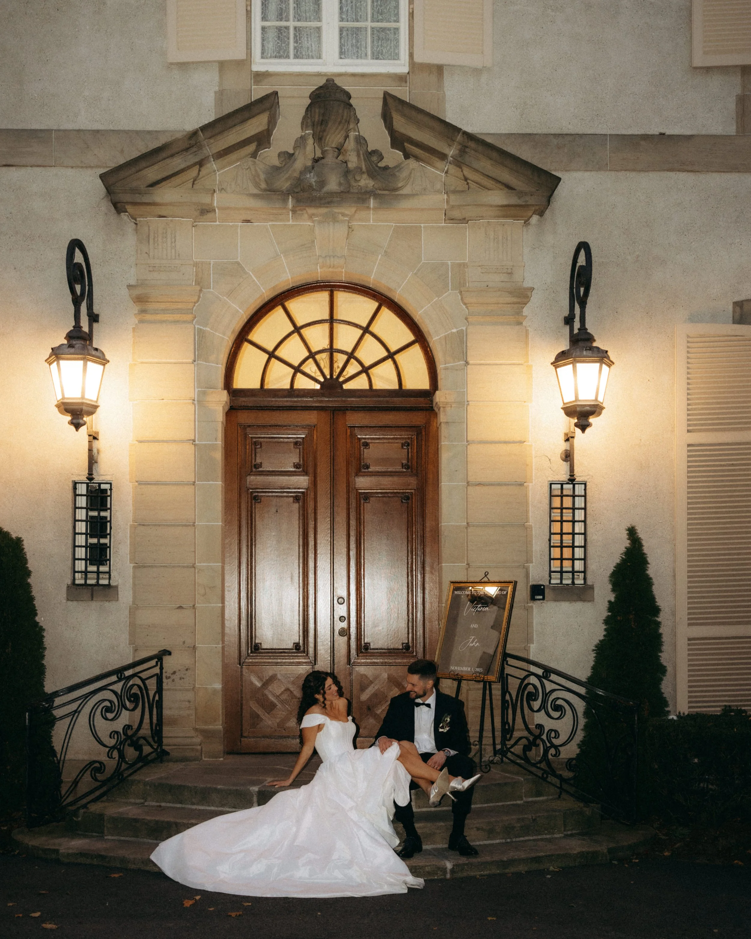 A bride and groom in wedding attire sitting on steps outside a building during nighttime. The bride is wearing a white gown and the groom in a tuxedo. They are looking at each other smiling, with a large wooden door, window, and decorative lights in 