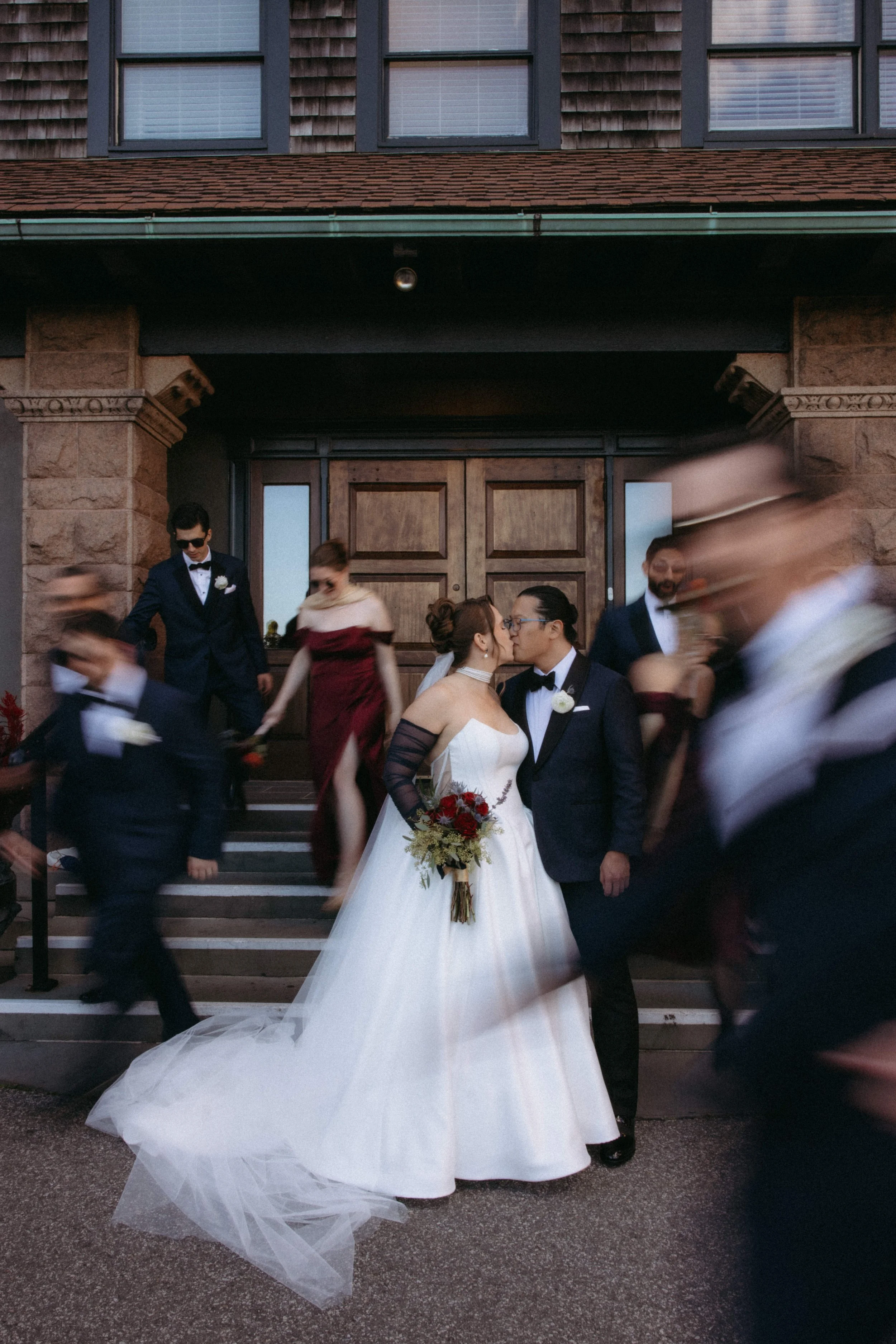 A bride and groom sharing a kiss on the steps in front of a building with large wooden doors, surrounded by wedding guests who are moving around.