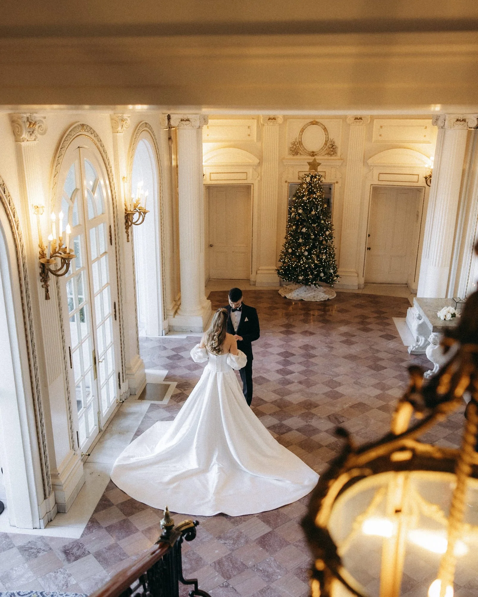 A bride and groom dancing in a grand, ornate hall decorated for Christmas with a Christmas tree. The hall features large windows, tall columns, and elegant lighting fixtures.