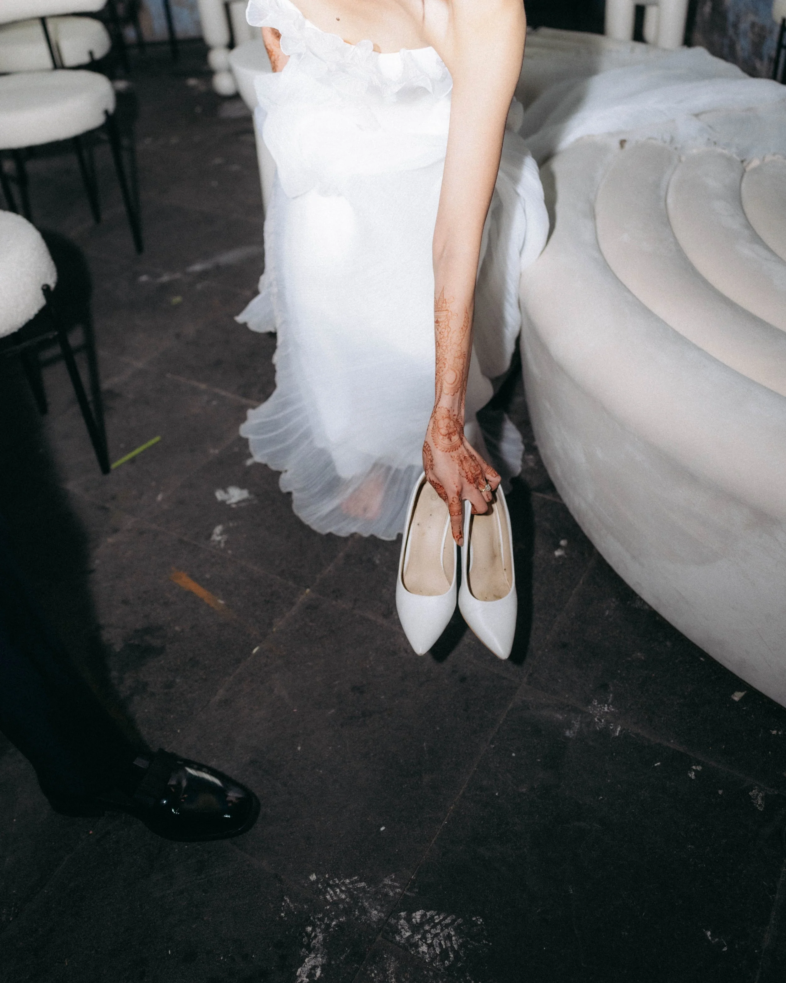 Person wearing a white dress with ruffles, holding white high-heeled shoes, with another pair of black shoes visible on the dark floor, in a room with white chairs and a white couch.