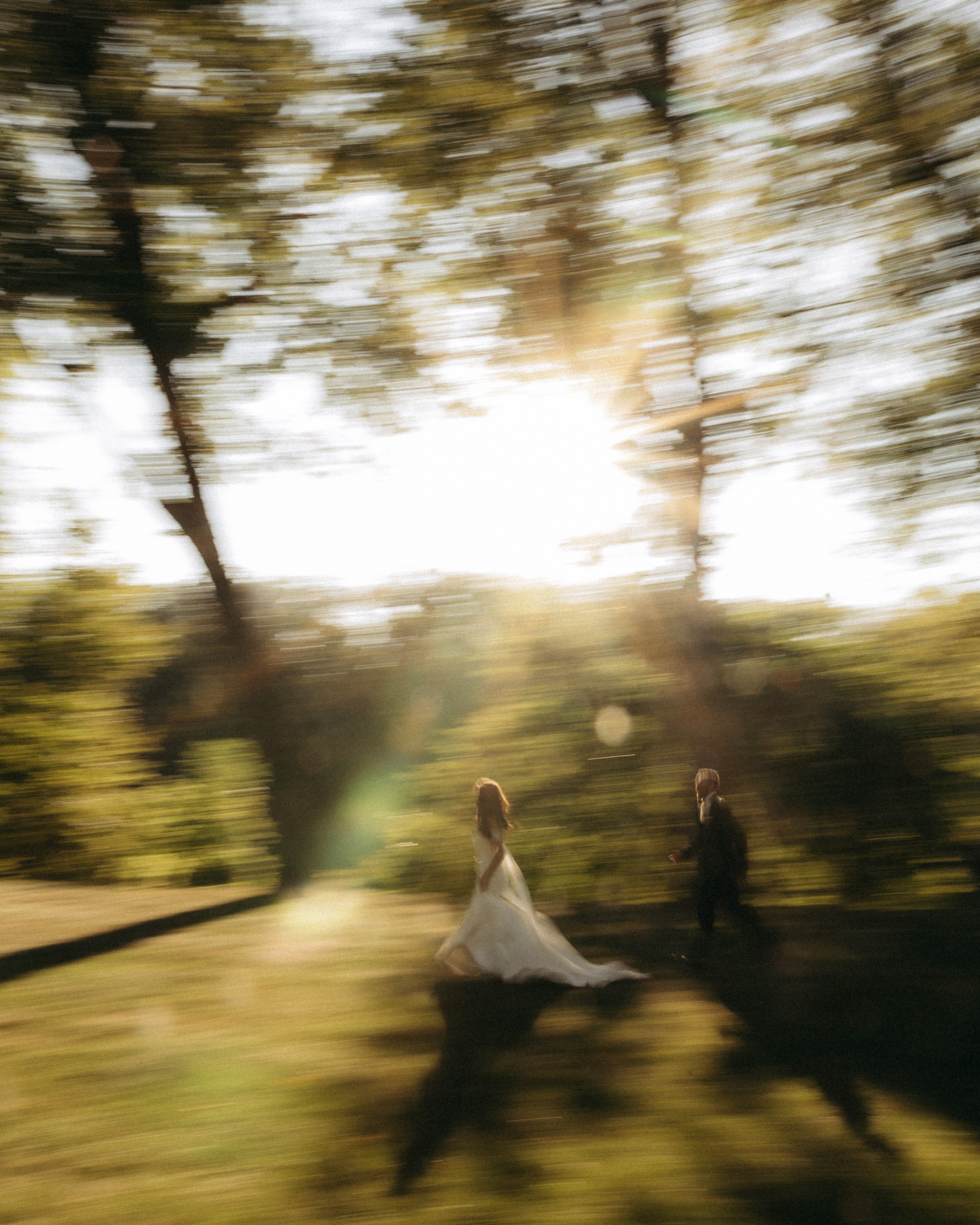 A blurred photo of a park with tall trees, taken during sunset. In the foreground, a bride in a white wedding gown and a groom in a dark suit are walking together.