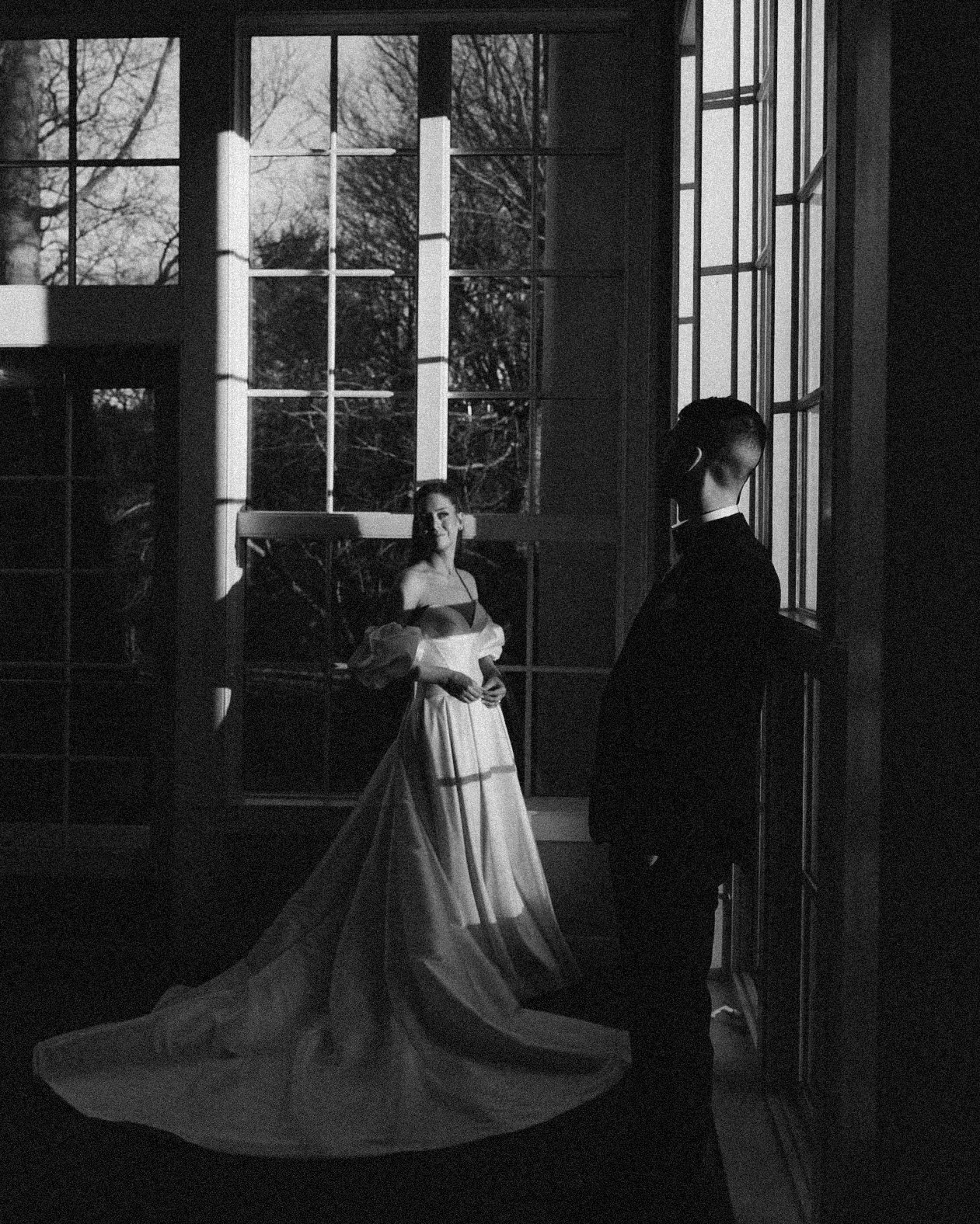 A black and white photograph of a woman in a long, flowing gown and a man in a tuxedo standing near large windows with natural light casting shadows inside. The woman is looking at the man, and the surroundings suggest a historic or elegant setting.