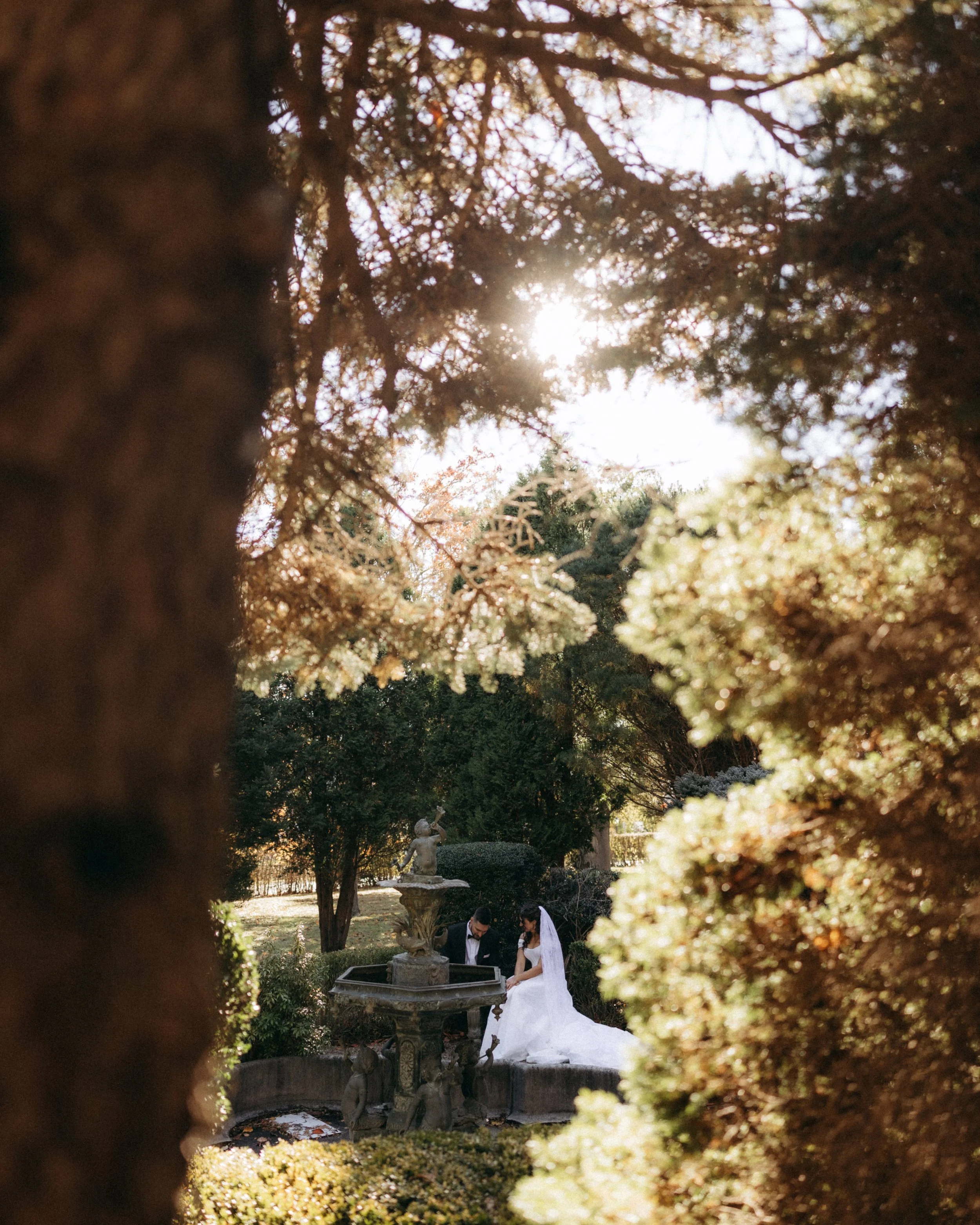 A bride and groom are sitting together near a fountain in a garden, framed by blurred foliage and branches with sunlight filtering through.